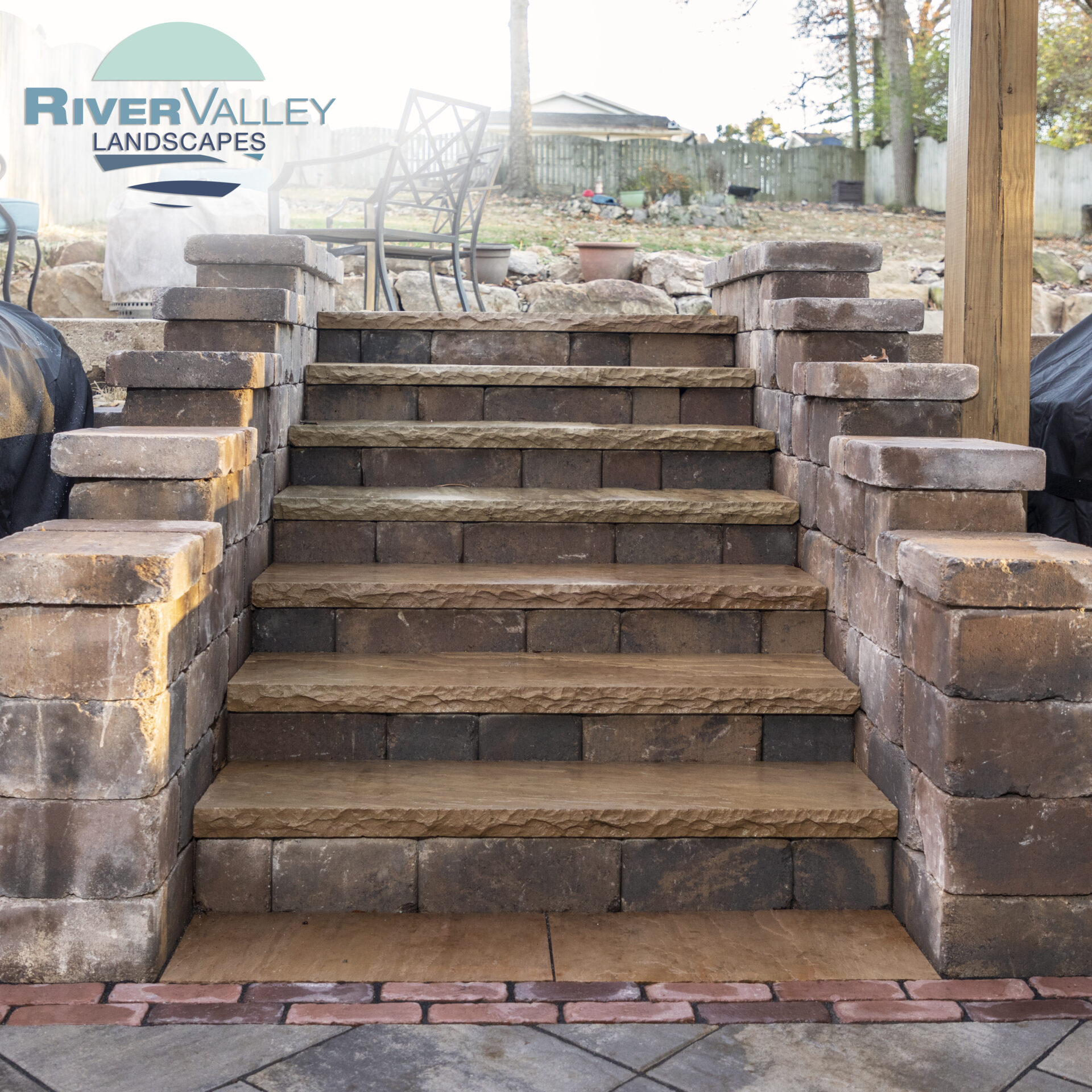 Stone steps with brick borders lead up to a seating area in a landscaped backyard, featuring a fence and patio furniture.