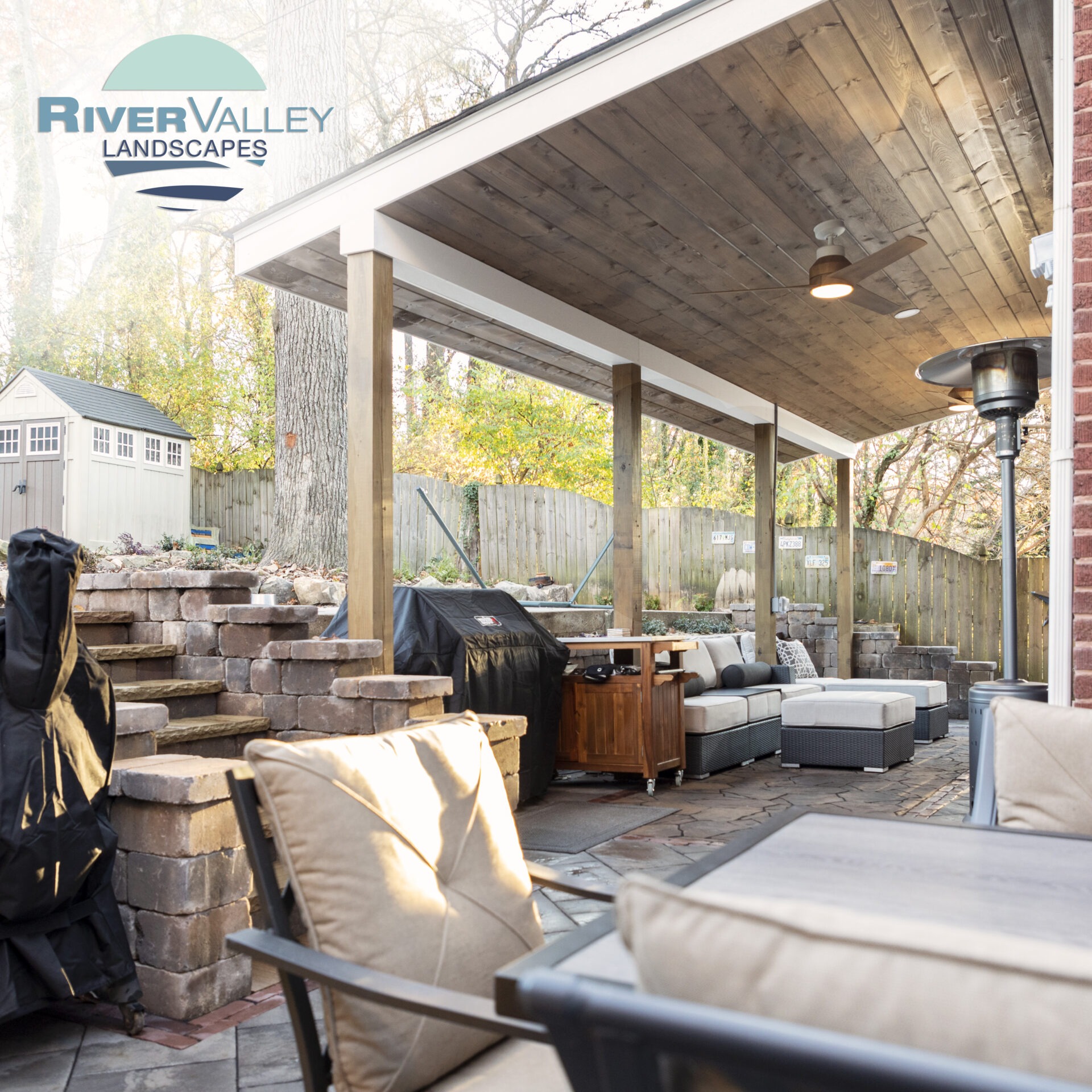 A cozy outdoor patio features cushioned seating, a wooden ceiling, barbecue grill, and heater, with a small shed and landscaped garden nearby.