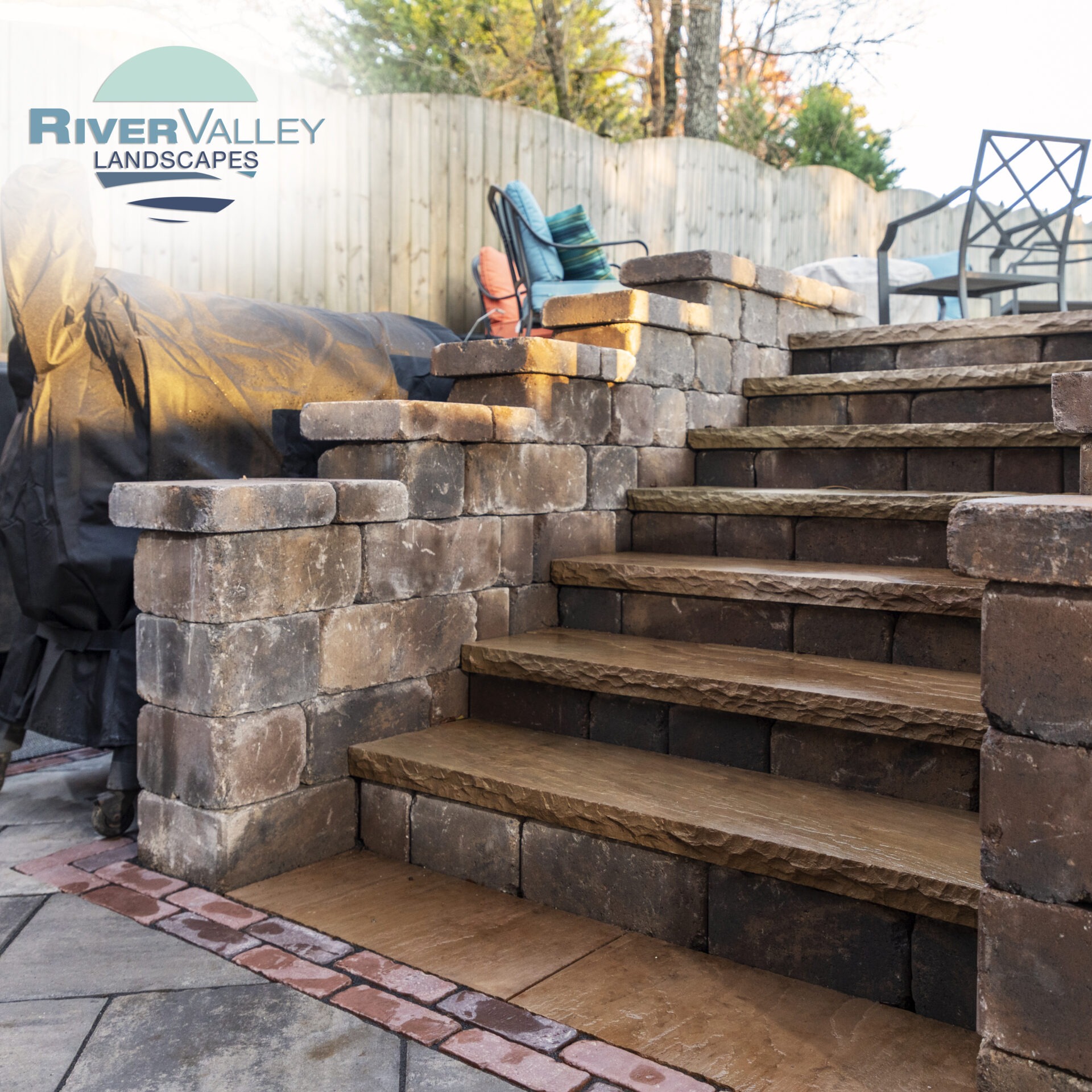 Rustic stone steps lead up to a landscaped patio with chairs and a covered grill. Sunlight highlights the outdoor seating area.