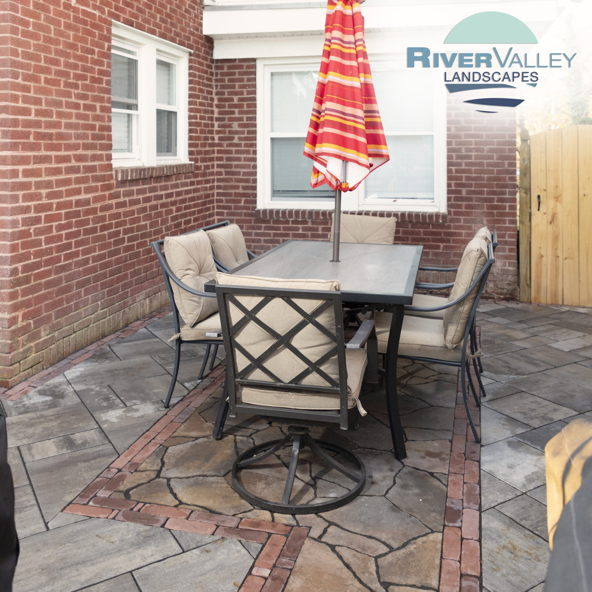 Outdoor patio with metal table, cushioned chairs, and a red-striped umbrella. Brick wall and patterned stone flooring create a cozy, enclosed space.