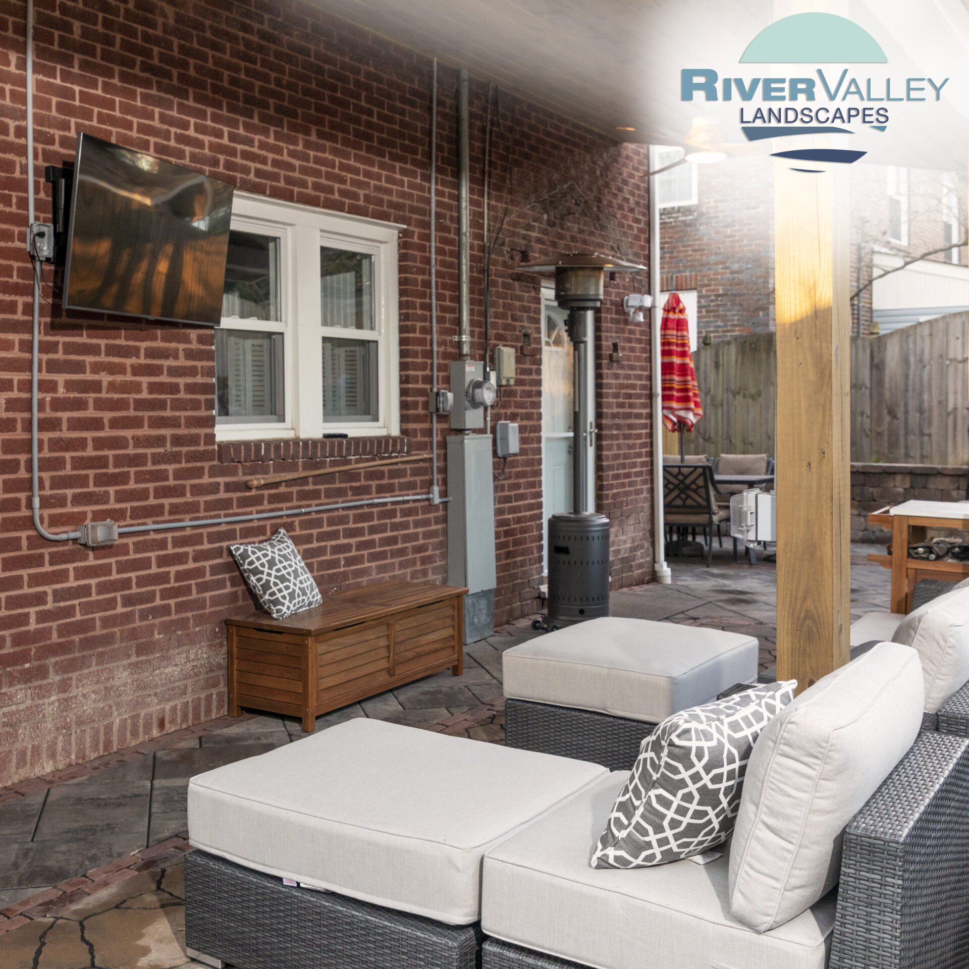 Cozy patio with modern furniture, TV, and heater against a brick wall. Logo of River Valley Landscapes in the top corner.