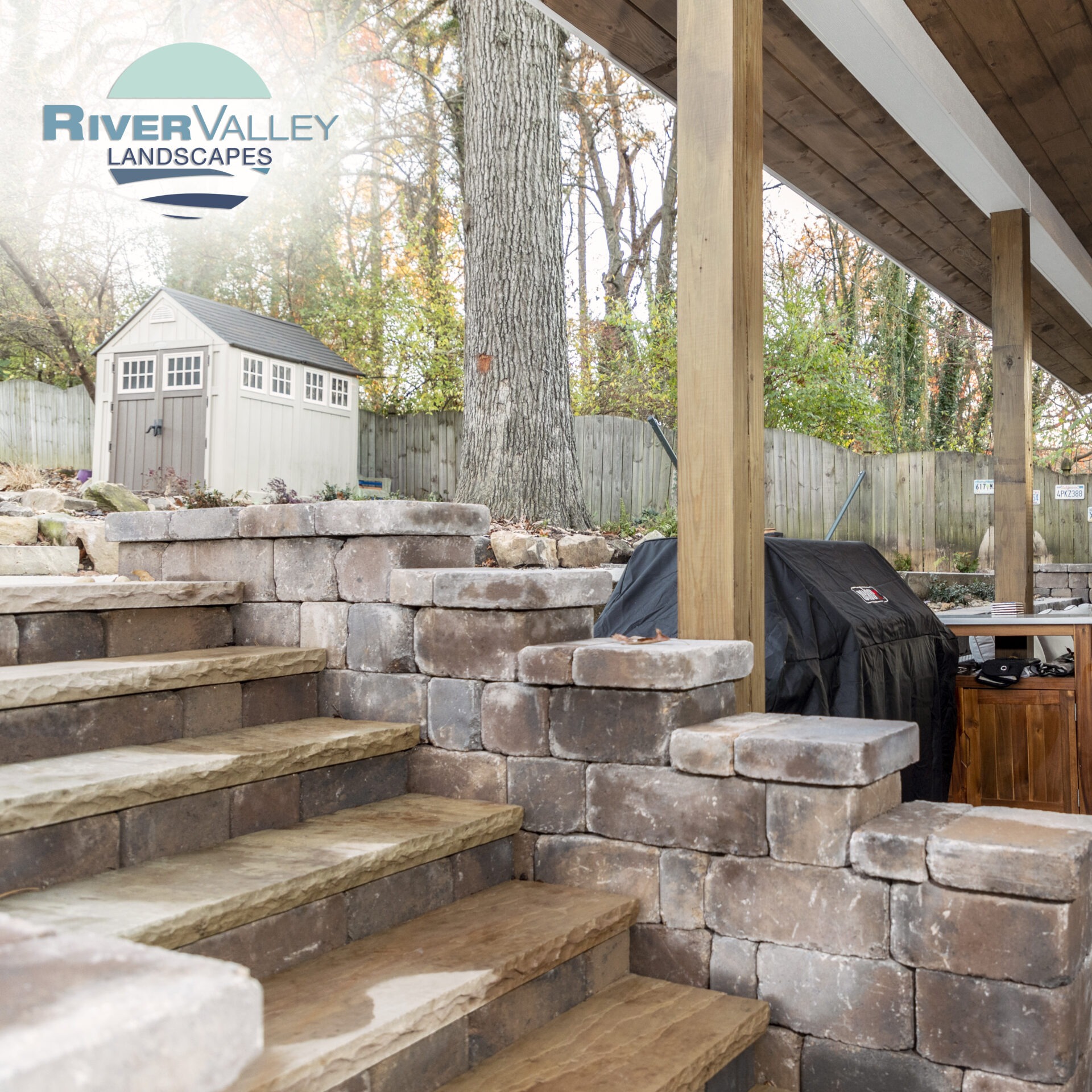 Stone steps leading to outdoor patio with covered grill, wooden posts, and a shed surrounded by trees and a fenced yard.