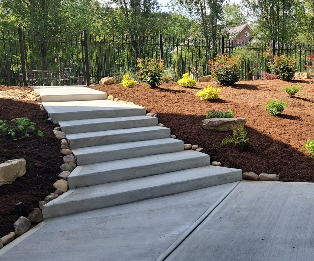 Concrete steps lead through landscaped garden with mulch, plants, and rocks, bordered by a metal fence. Trees and a house in background.