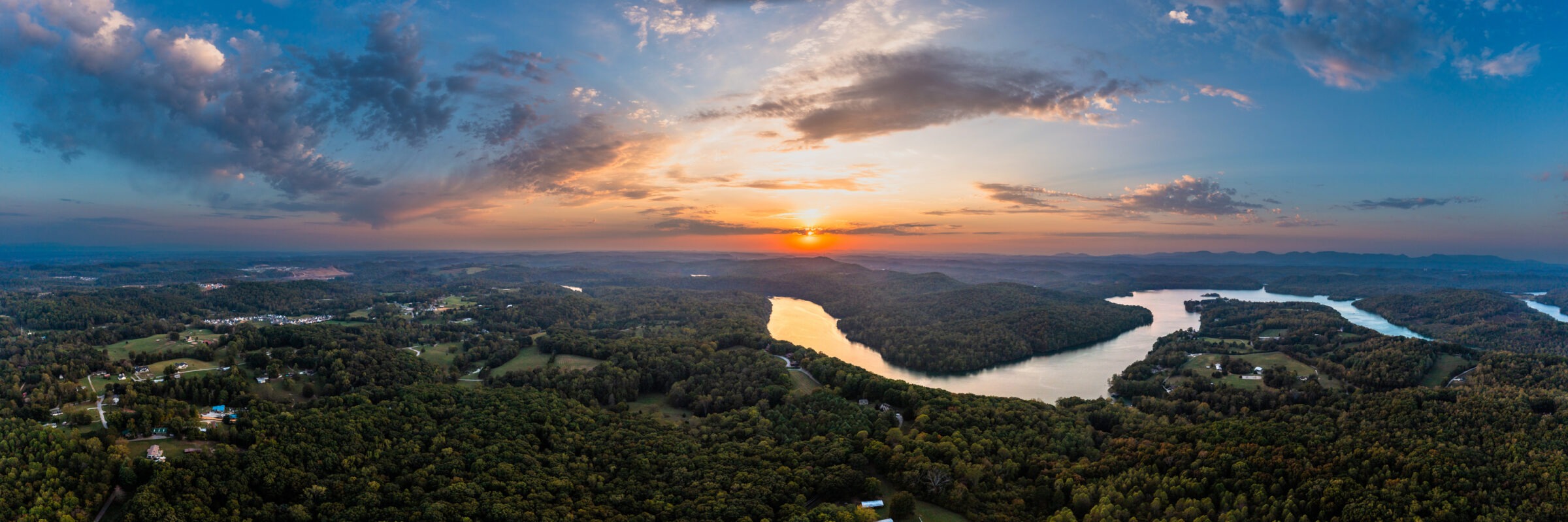 Aerial view of a serene lake at sunset, surrounded by lush forests and scattered houses, under a dramatic, colorful sky.