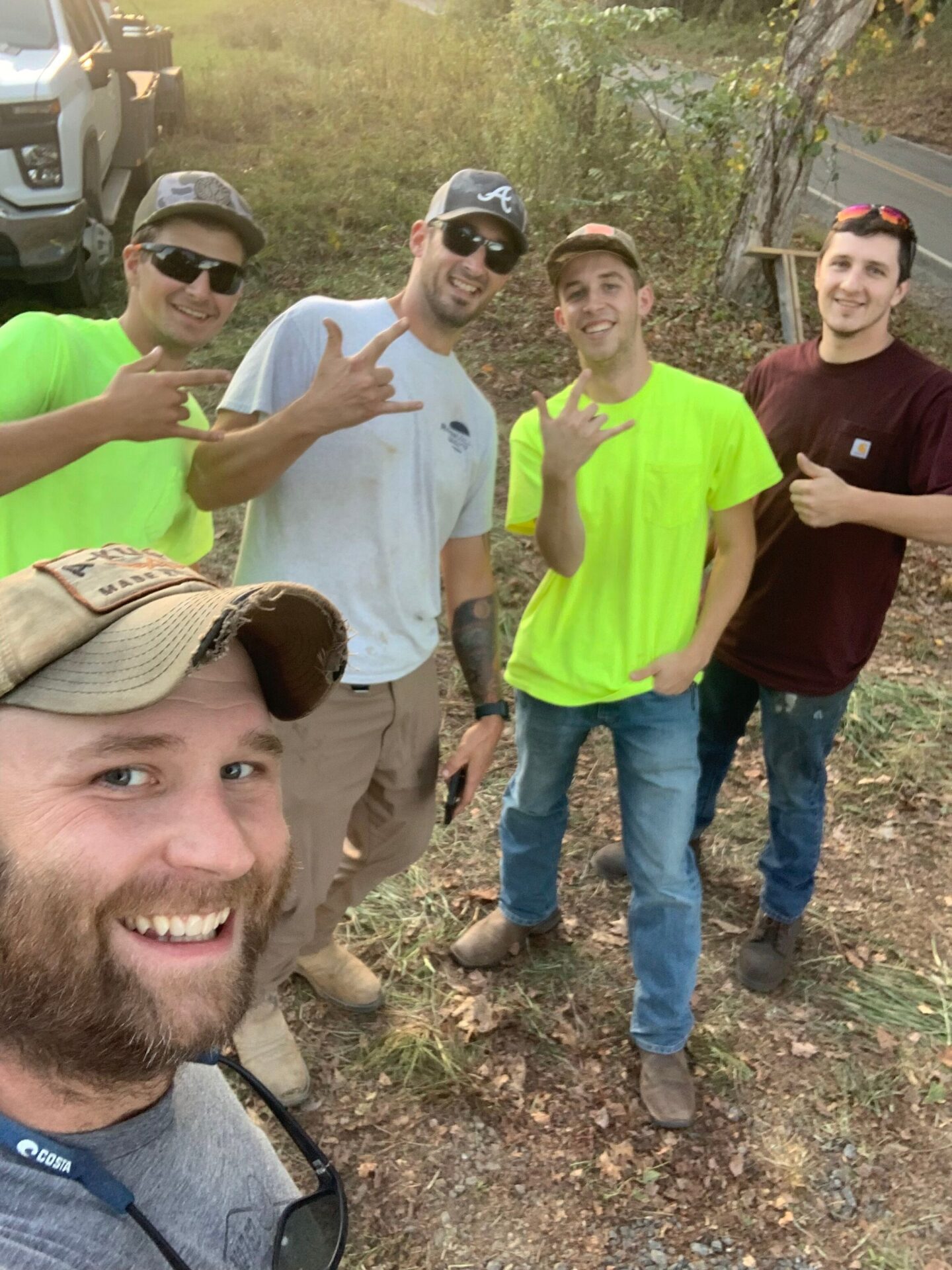 Five people outdoors, smiling and gesturing at the camera. They're near a road, surrounded by grass and trees, with a truck in view.