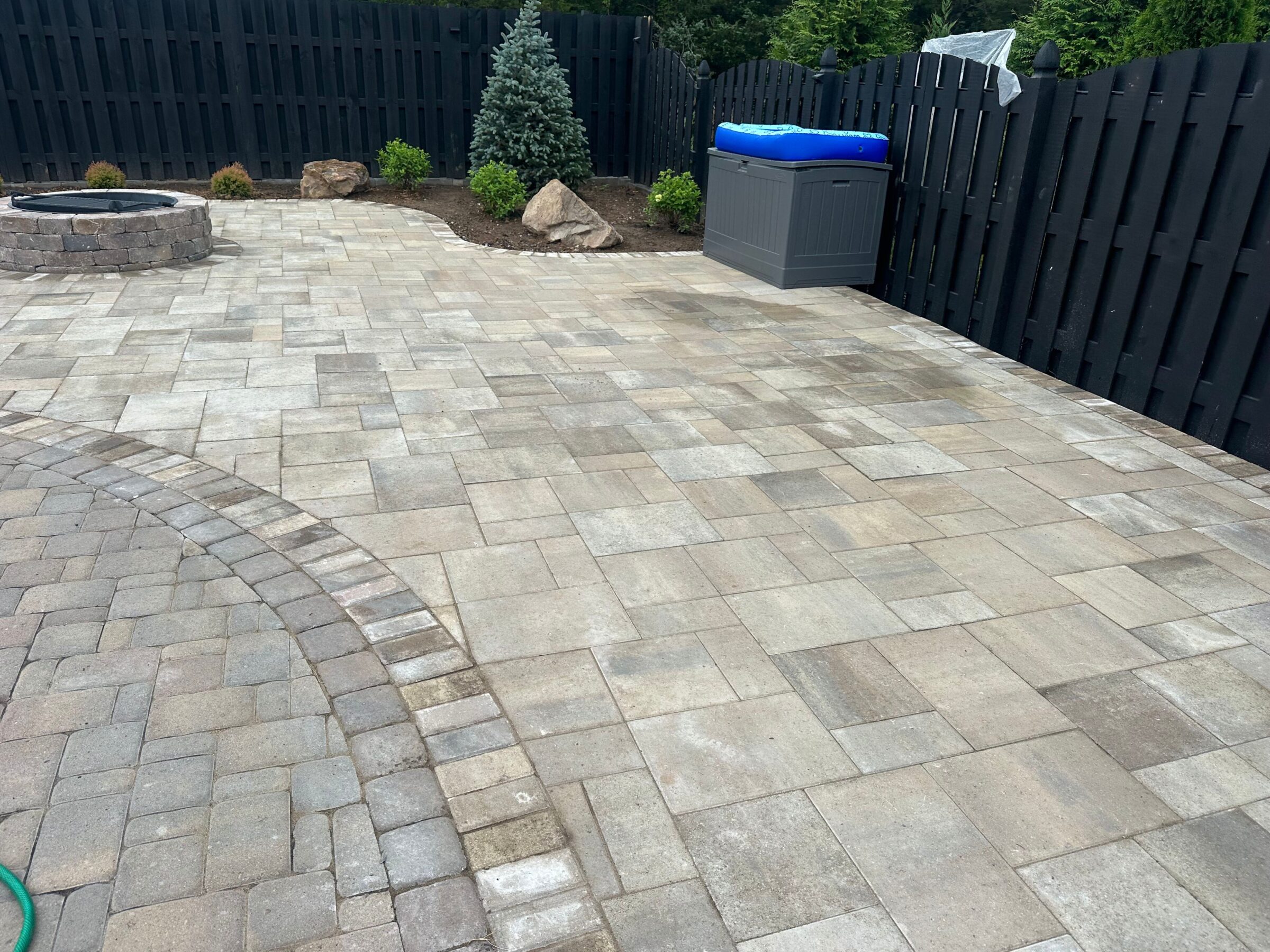 A spacious backyard patio with geometric paving stones, a round fire pit, small plants, and a black wooden fence with a storage box.