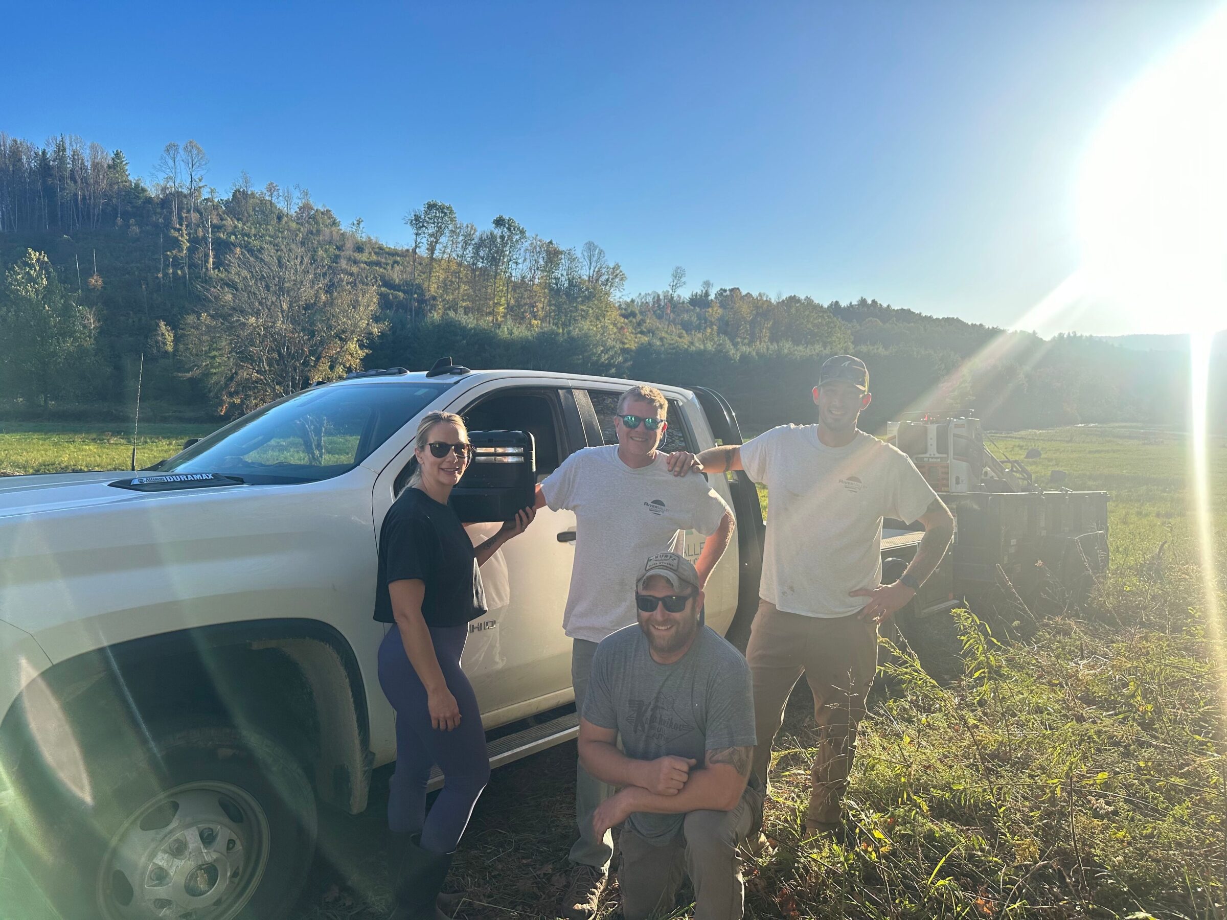 Four people pose beside a white truck in a sunlit, grassy field with wooded hills in the background.