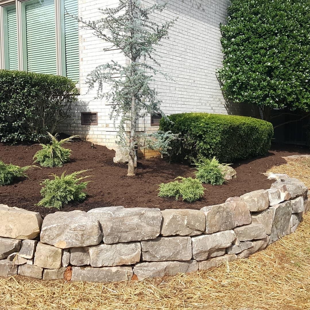 A landscaped garden with shrubs and a small tree is bordered by a stone wall against a light brick building exterior.