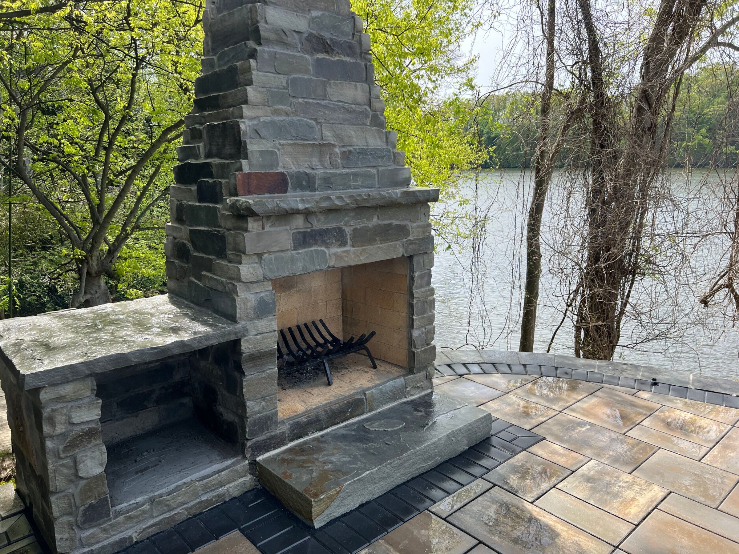 Stone outdoor fireplace on a spacious patio overlooking a tranquil river, surrounded by lush green trees in a serene natural setting.