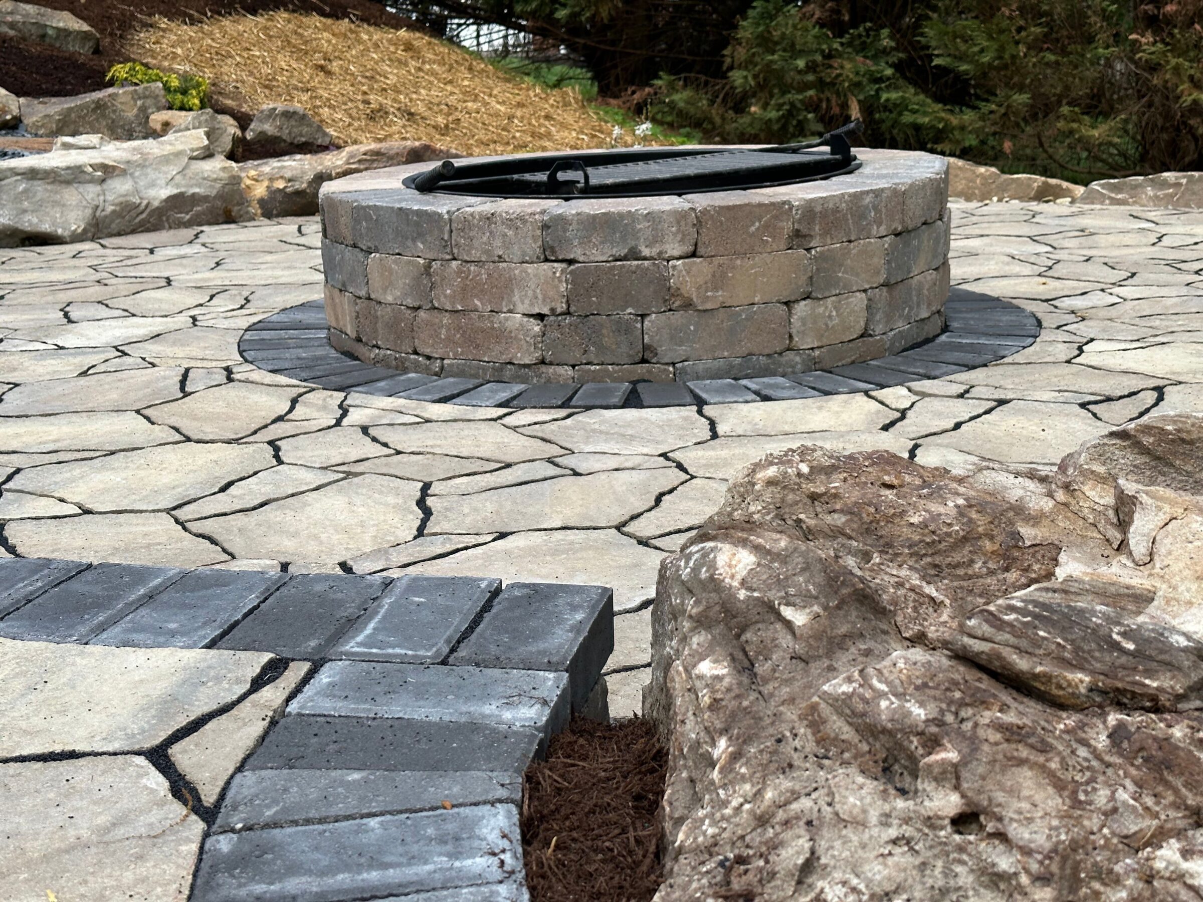 A circular stone fire pit surrounded by a patterned patio with natural rocks, set in an outdoor garden area.