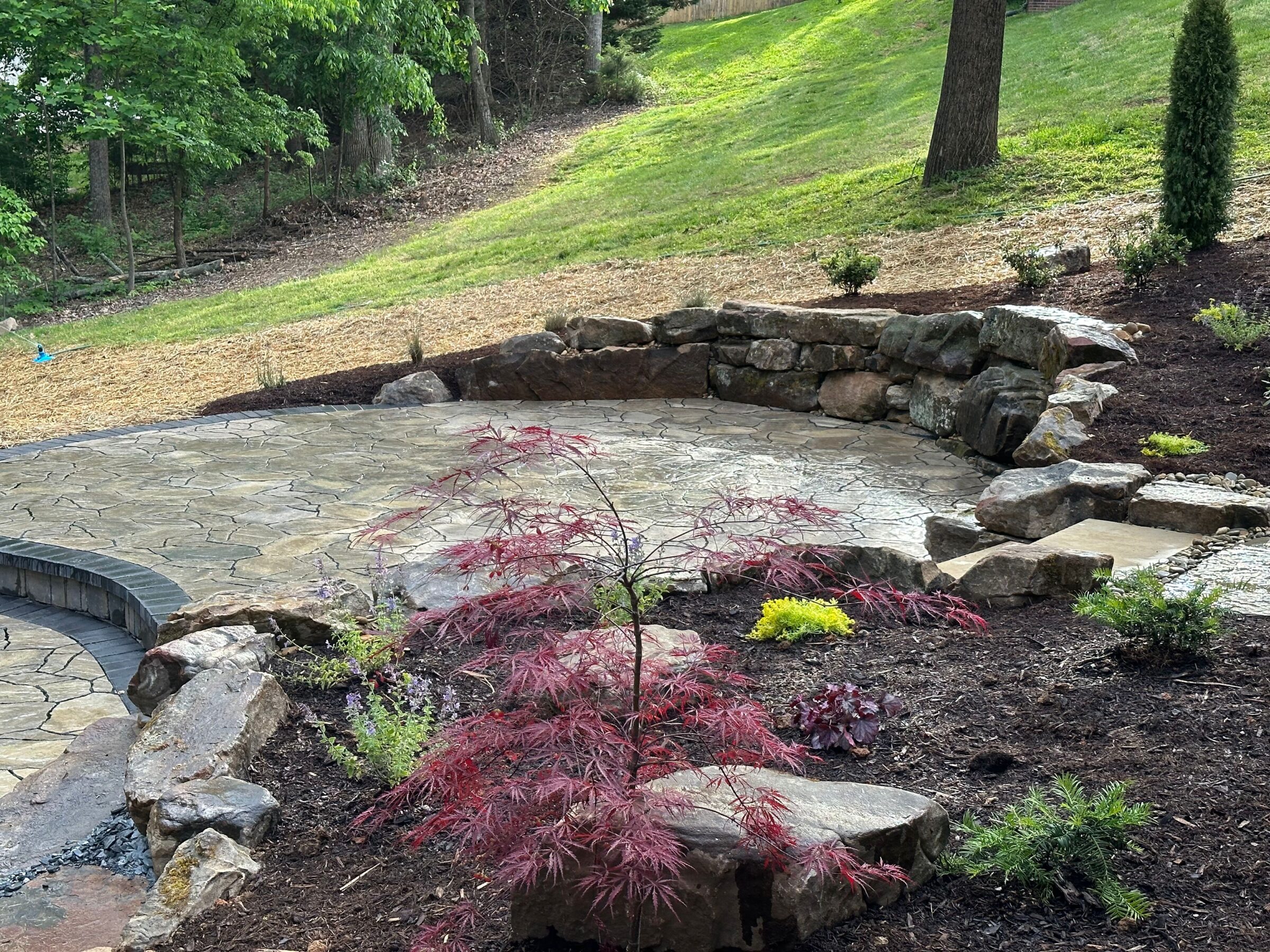 A landscaped garden features a stone patio surrounded by rocks, various plants, and trees with a grassy slope in the background.