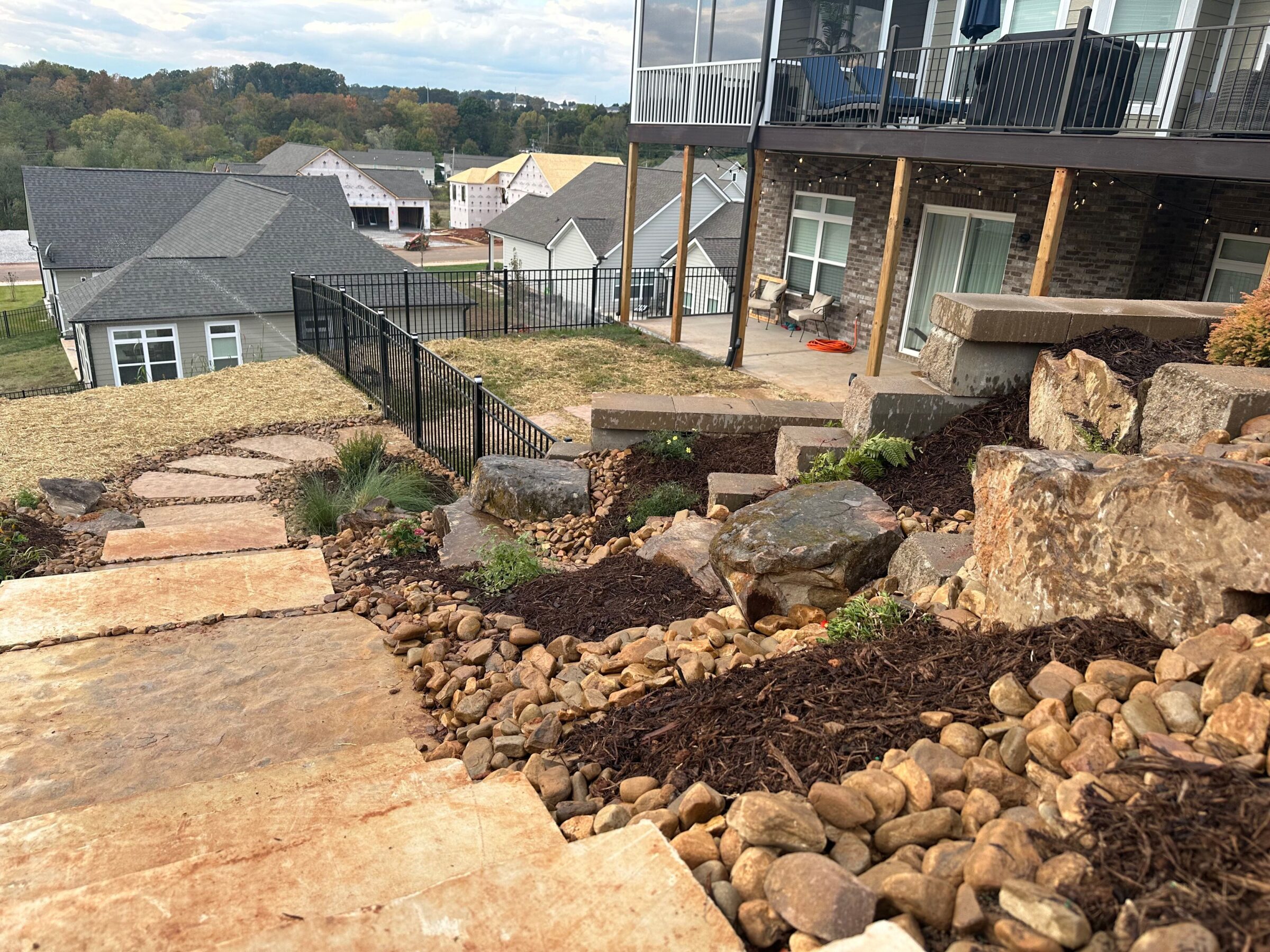 A landscaped backyard features stone steps and rock garden. Houses and a patio with furniture overlook a scenic, wooded backdrop under a cloudy sky.