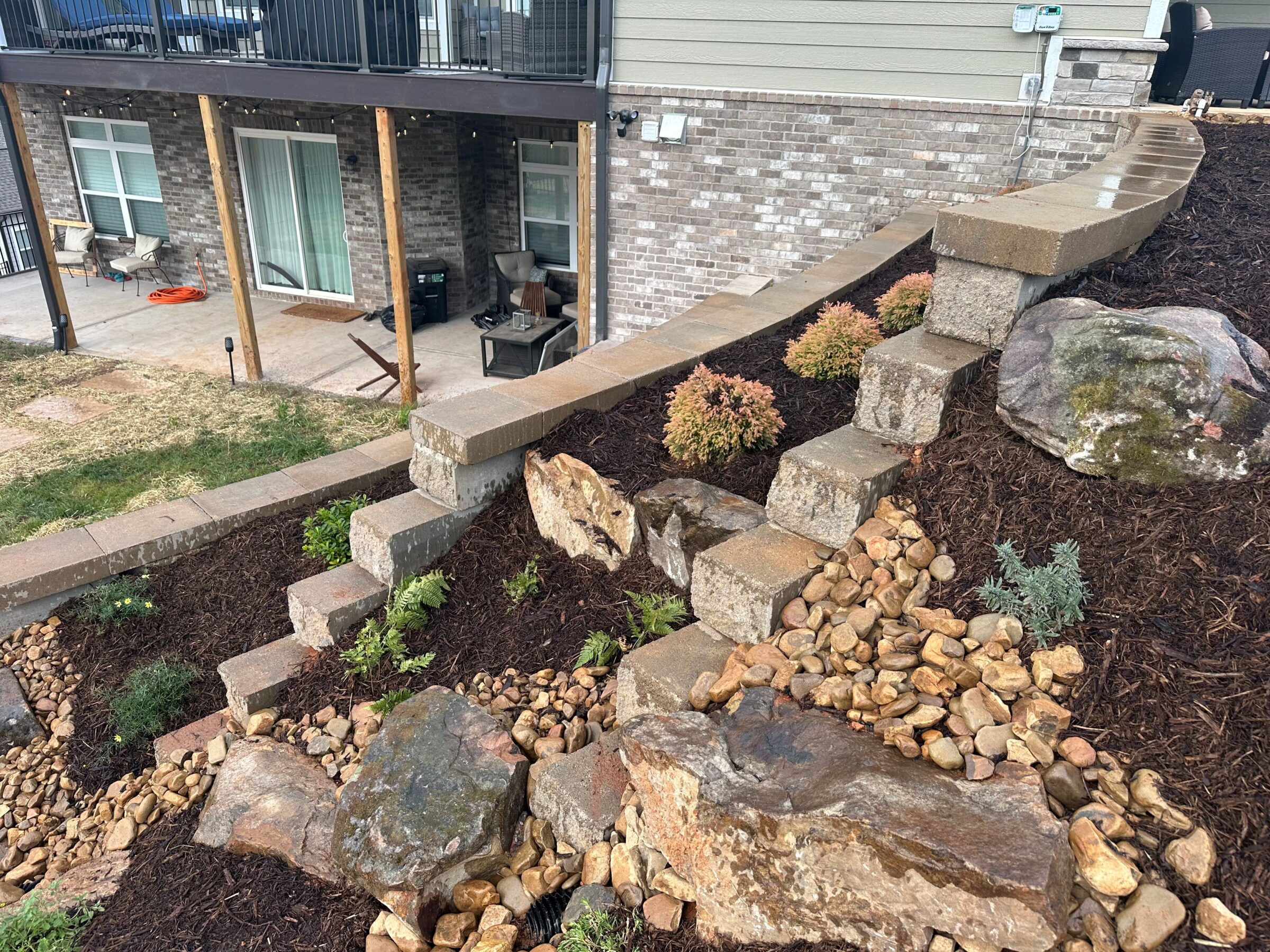 Stone steps lead through a landscaped garden with rocks and plants beside a brick house porch with outdoor seating.