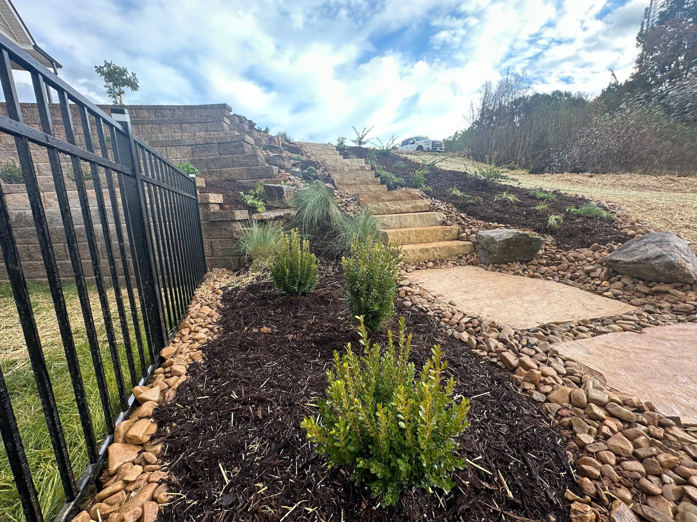 A landscaped garden with stone steps, bordered by mulch, plants, and rocks, ascends a hill beside a railing under a partly cloudy sky.