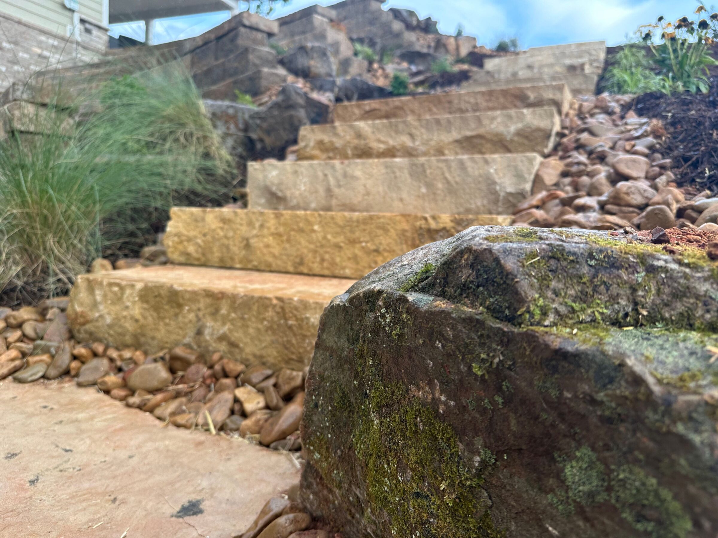 Stone steps ascend amidst rocks and greenery in a natural garden setting under a clear sky.
