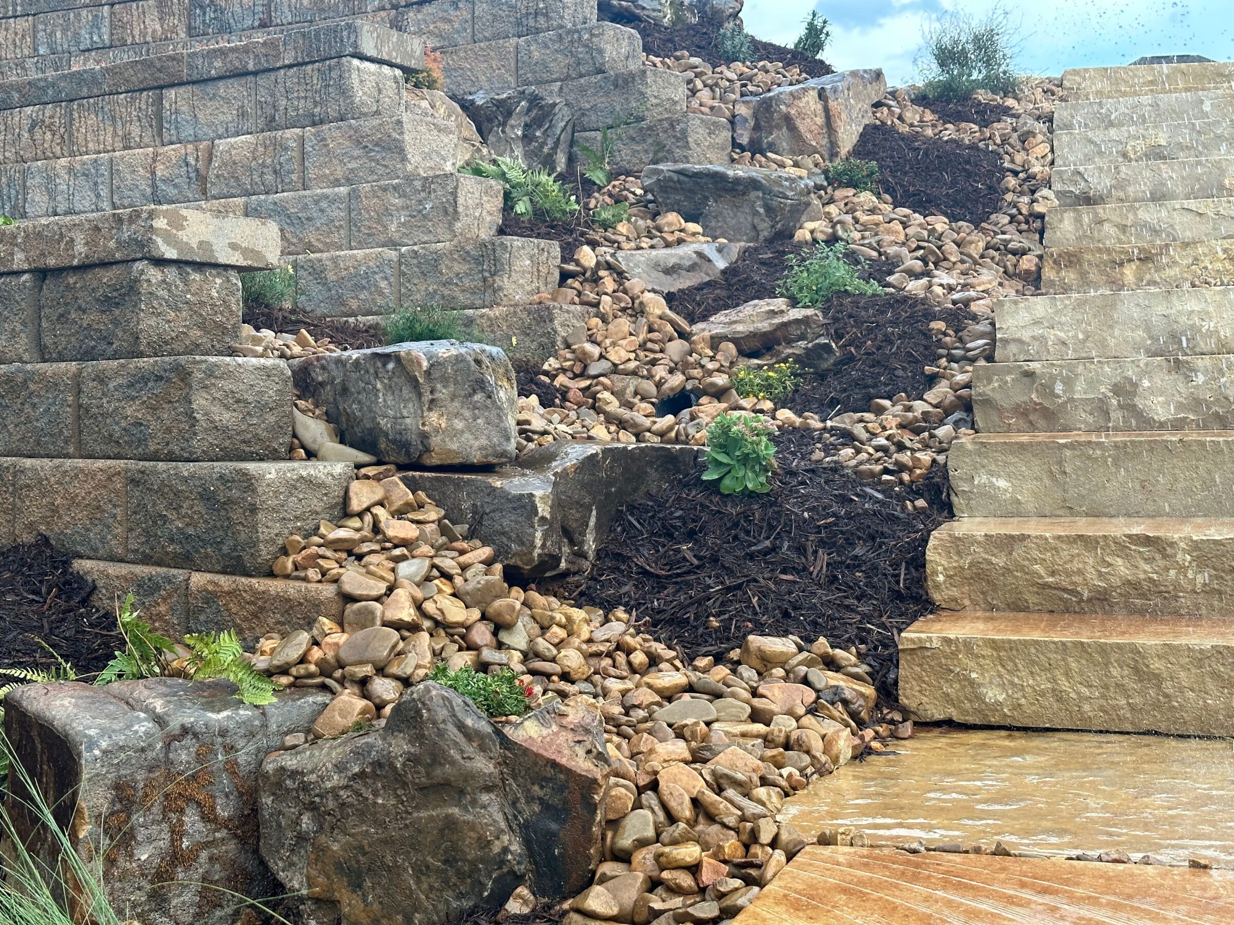 Stone steps and retaining wall with decorative rocks, plants, and mulch, create a natural landscape design under a partly cloudy sky.