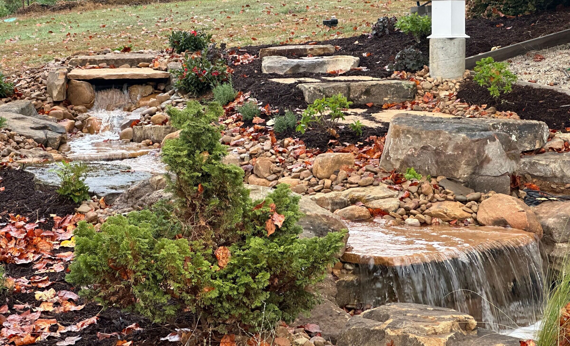 A serene garden with a small cascading waterfall, surrounded by rocks, plants, and autumn leaves, exuding a peaceful, natural ambiance.