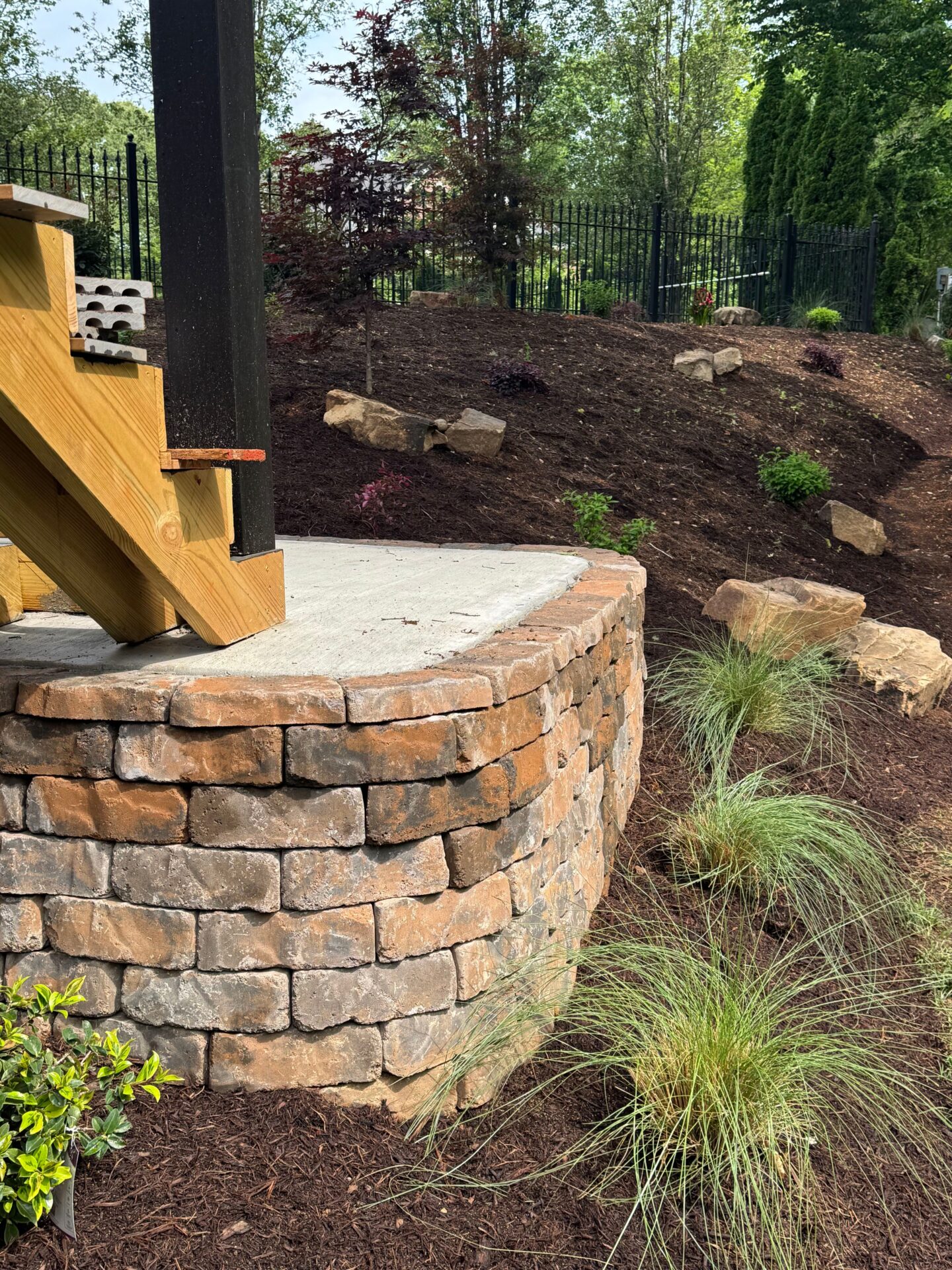 A landscaped garden features stacked stone wall and steps with ornamental plants, tall grasses, and a black metal fence in the background.