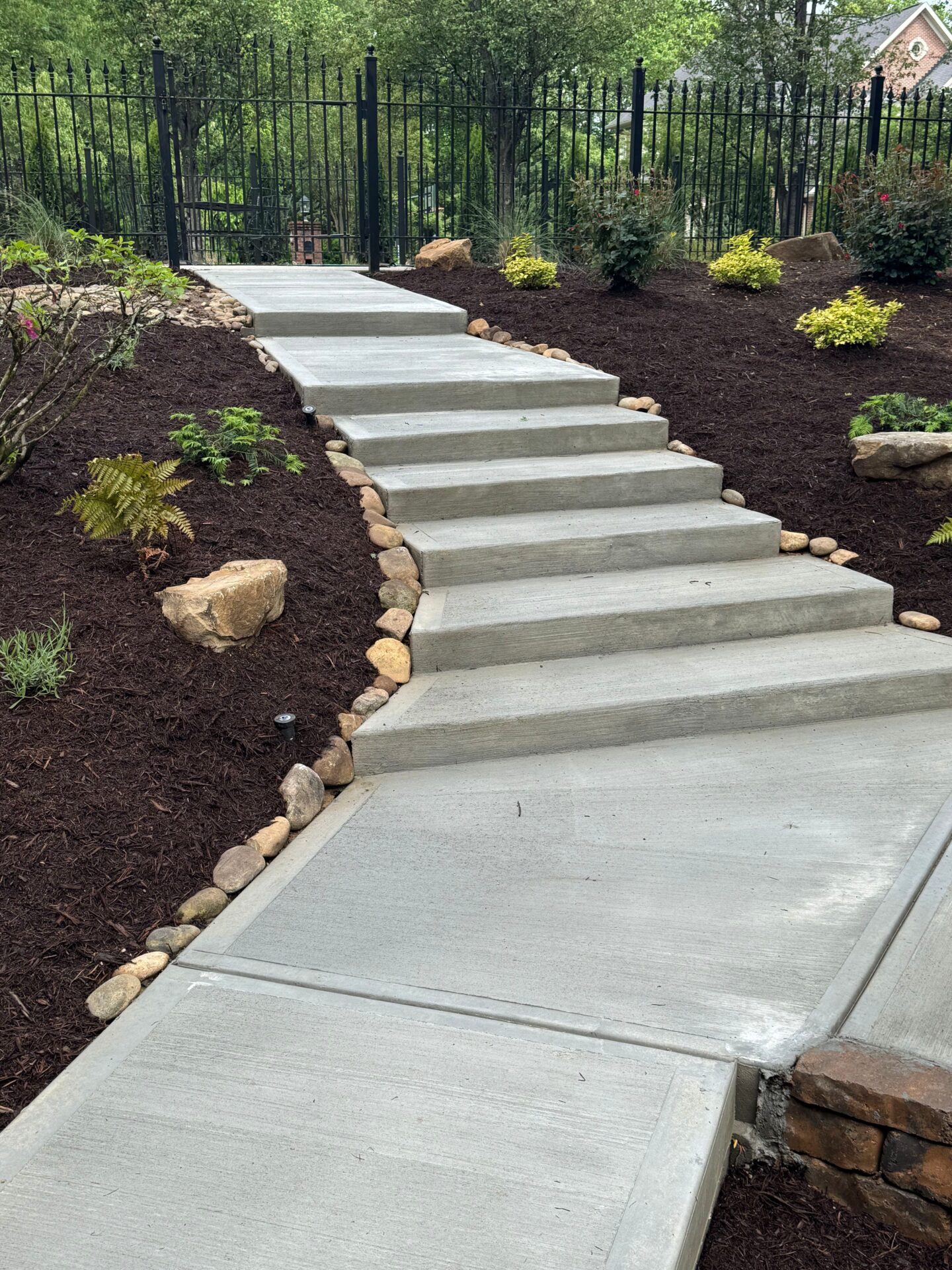 Concrete steps ascend through landscaped garden, lined with rocks and bordered by black iron fencing, leading towards trees and residential area.