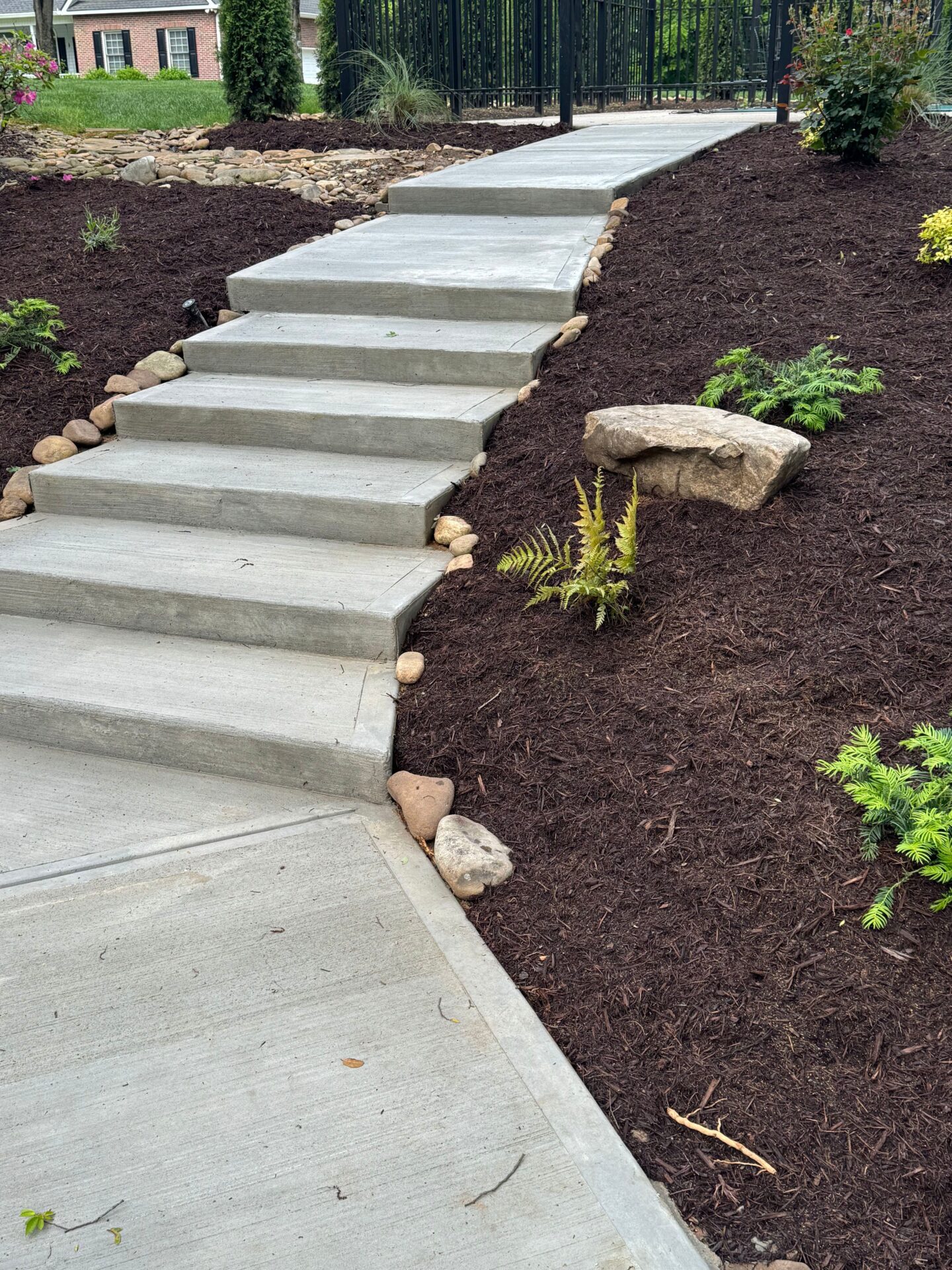 Concrete steps lead up through landscaped garden with mulch, rocks, and plants, framed by a metal fence; a house is visible beyond.