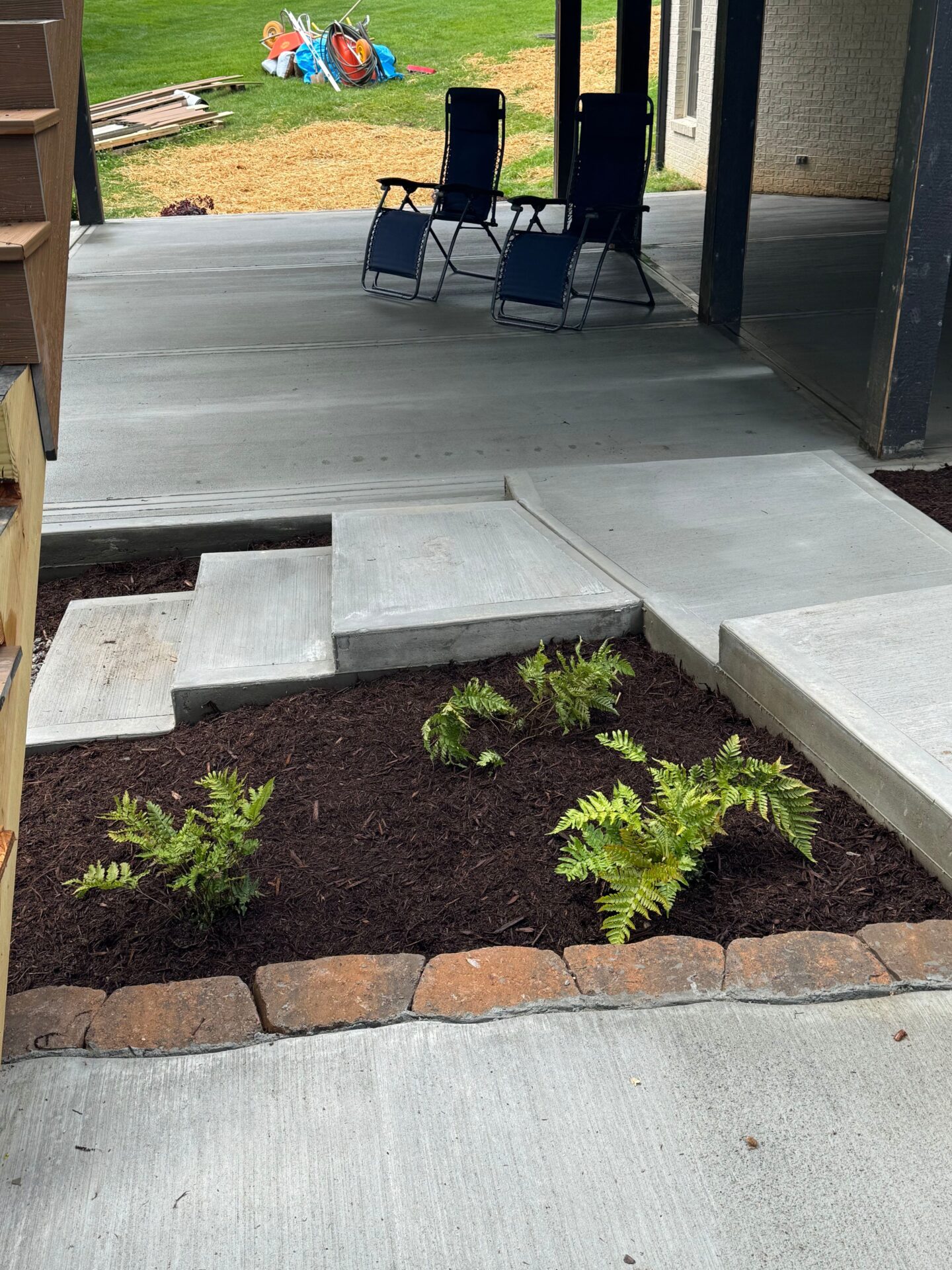 Concrete patio with two chairs, landscaped area with ferns. Background features lawn with scattered tools and supplies, including a wheelbarrow and tarps.