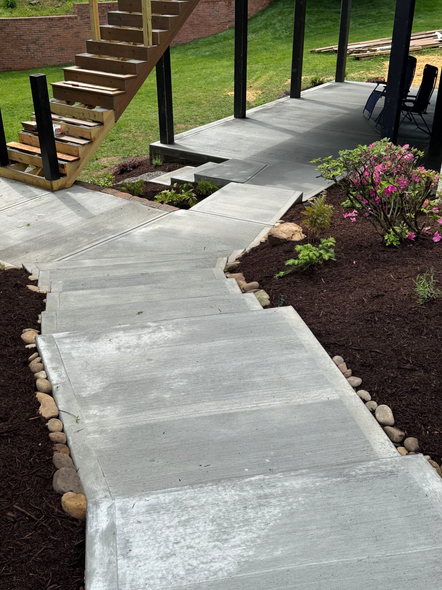 A newly constructed concrete walkway leads to stairs, surrounded by flowering bushes and mulch, with a patio area visible in the background.