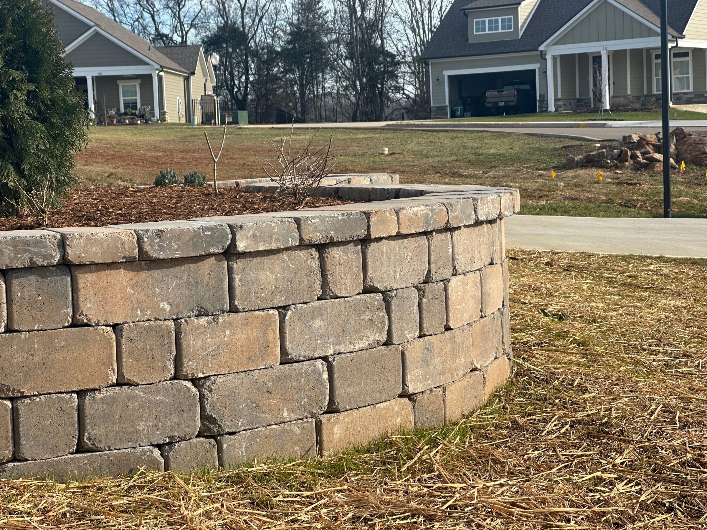 The image shows a stone retaining wall with suburban homes in the background under a clear sky, surrounded by bare trees and grass.