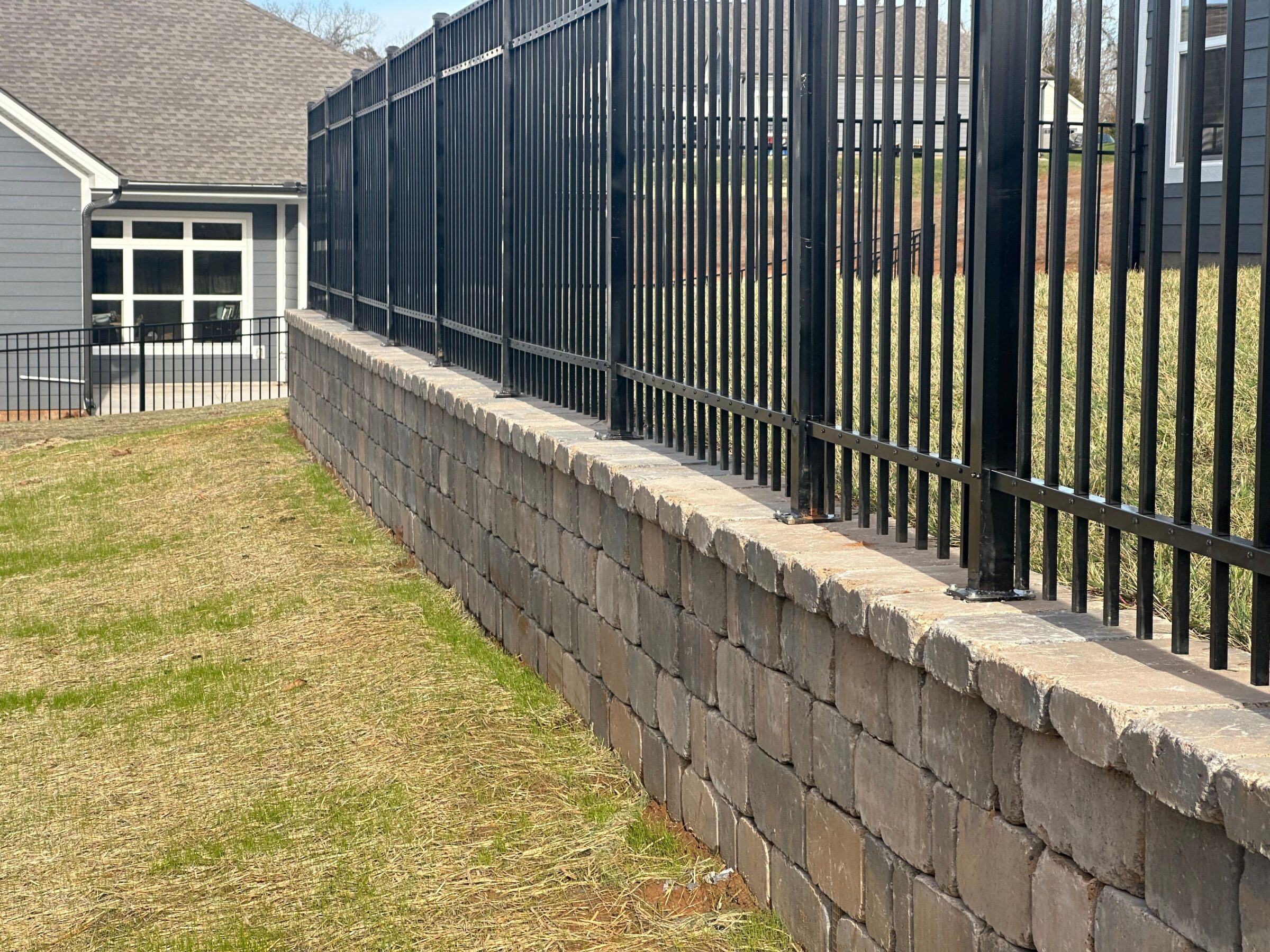 A brick retaining wall with a black metal fence borders a residential lawn, adjacent to modern houses and neatly trimmed grass.