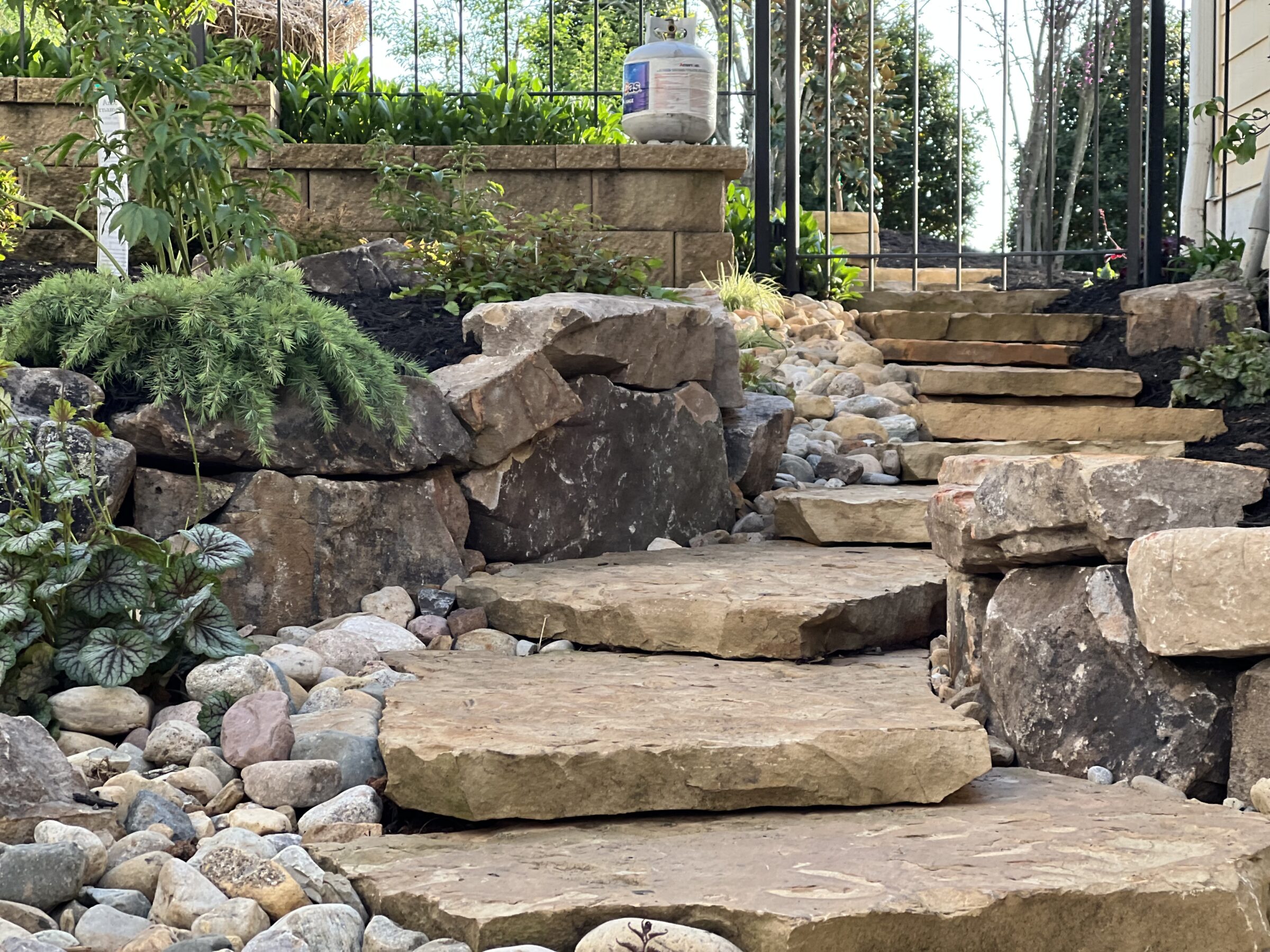 A stone pathway and steps are surrounded by rocks and lush greenery, leading up to a wrought iron gate with plants.