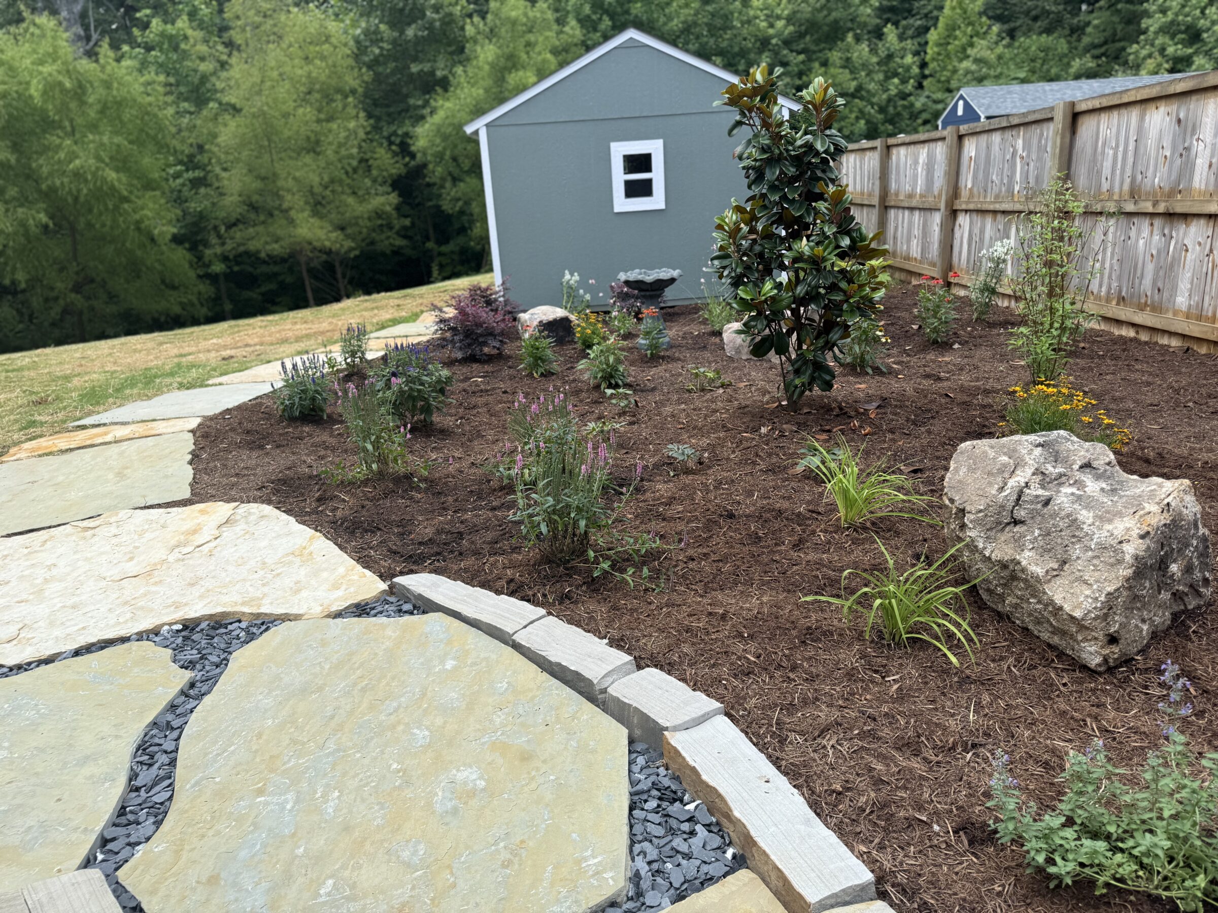 A well-maintained garden features a paved stone path, mulched plant beds, small green shed, and wooden fence surrounded by lush trees.