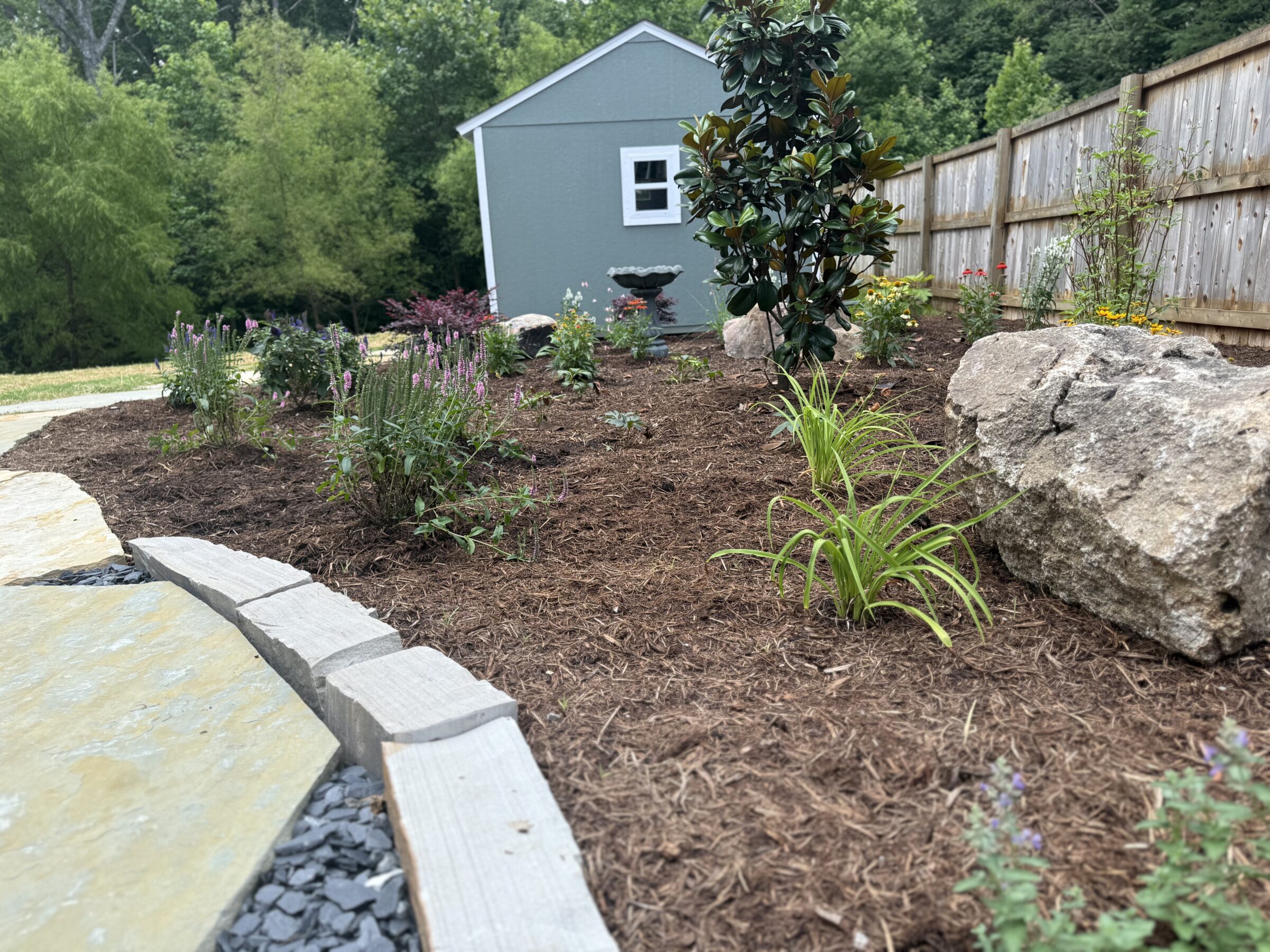 A landscaped garden with mulch, flowers, rocks, and a wooden fence. A small gray shed is in the background amidst greenery.