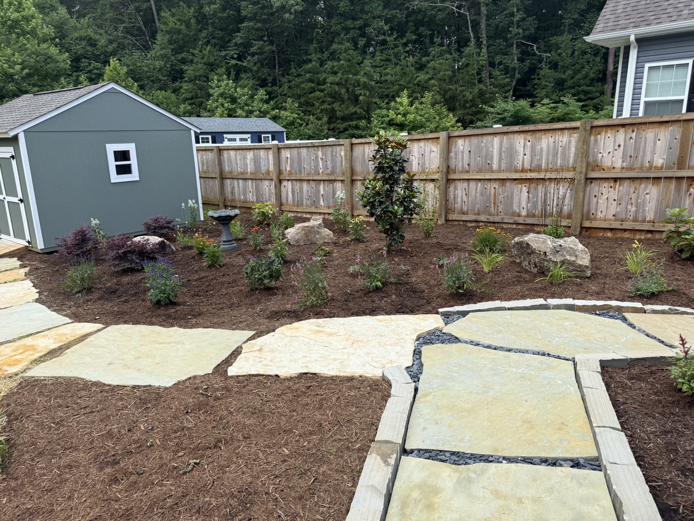 A landscaped backyard features a stone pathway, a wooden fence, two small sheds, and assorted plants and rocks surrounded by mulch.
