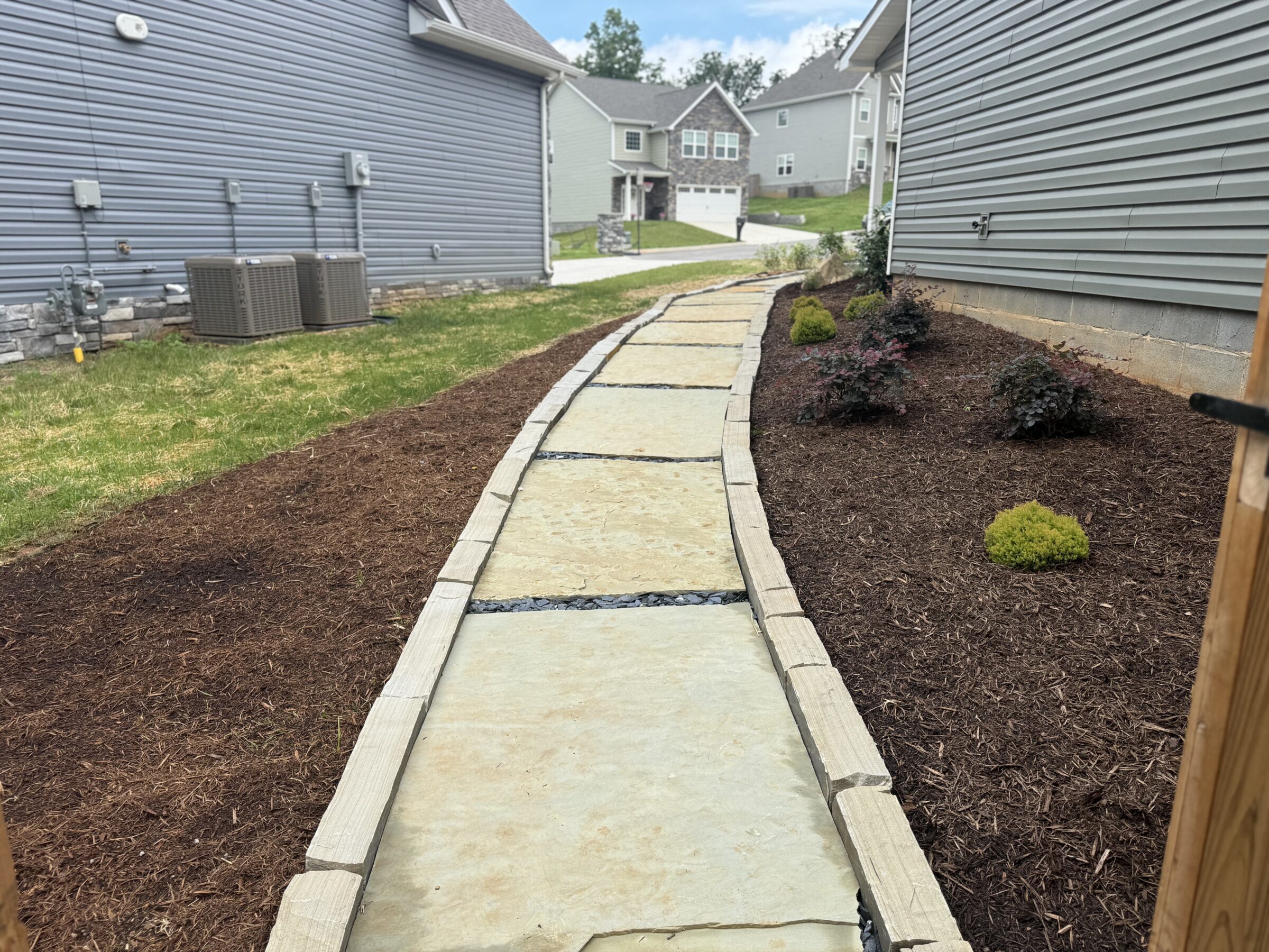 A stone pathway runs between two modern houses, bordered by mulch and small plants, leading towards more homes in the background.
