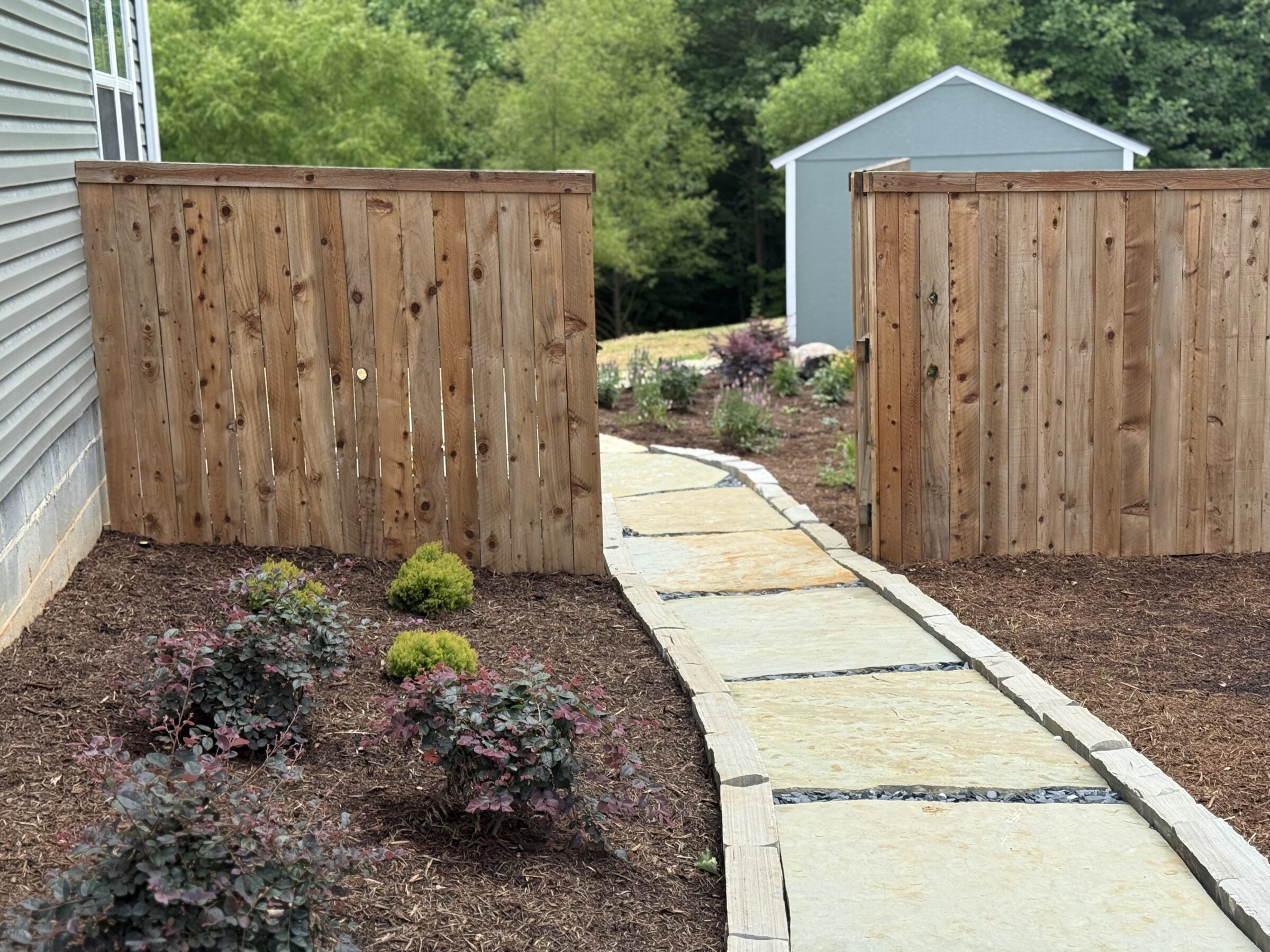 A stone path leads through wooden gates to a small shed, surrounded by planted shrubs and trees, under a clear sky.