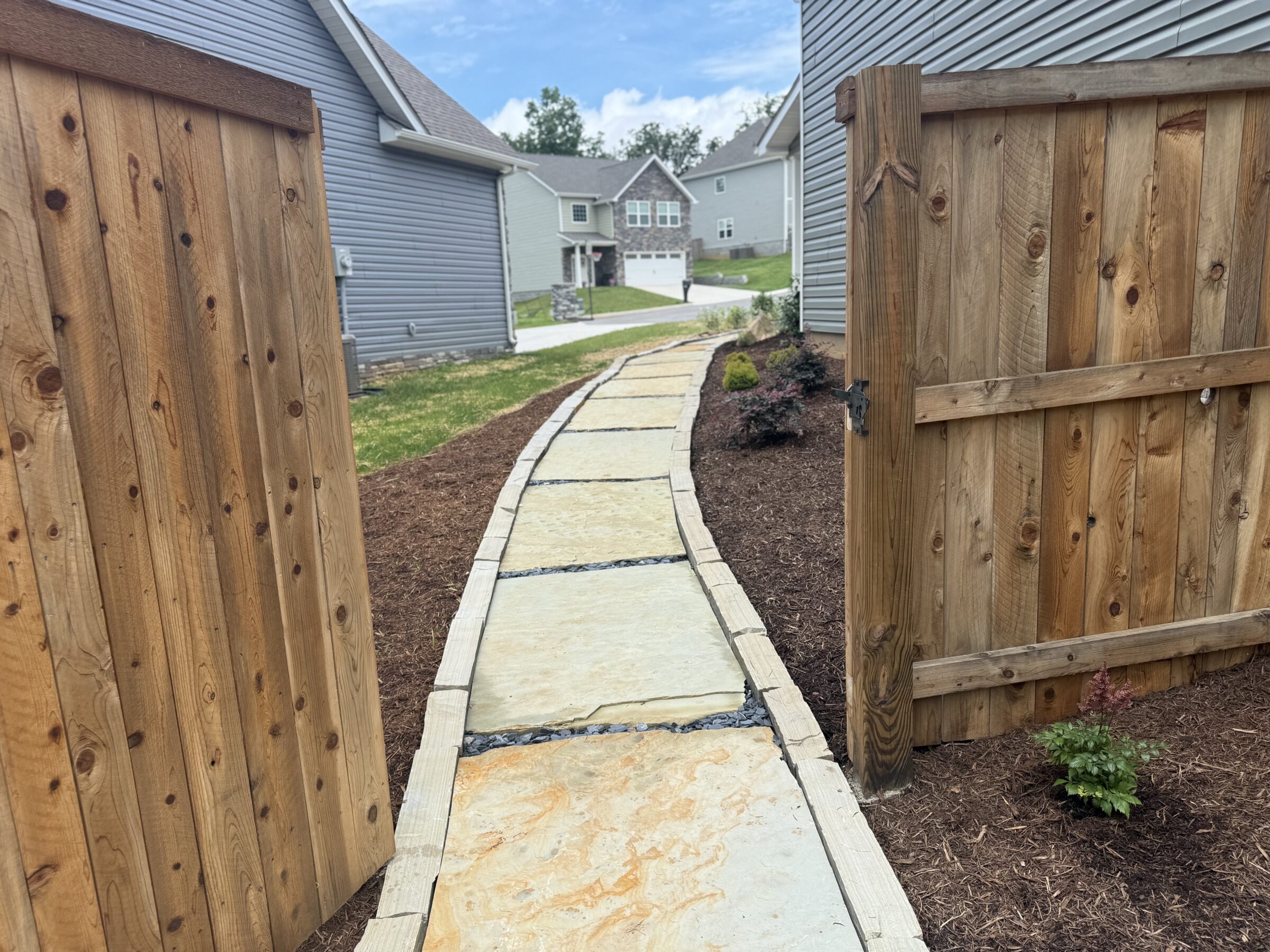 Wooden gates open to a stone path beside modern suburban houses. Mulched garden beds and a bright blue sky complete the scene.