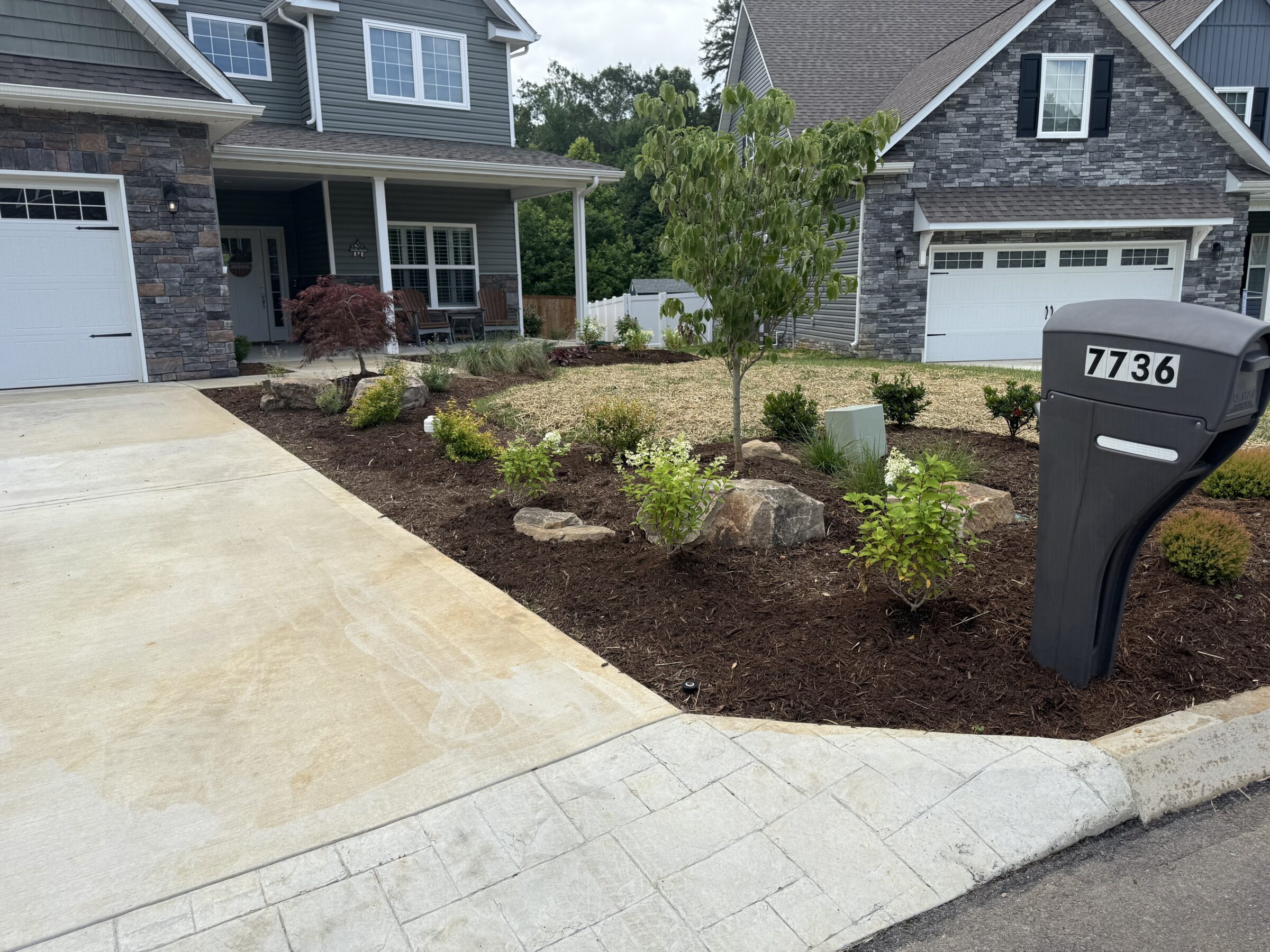 A modern two-story house with stone facade, landscaped front yard, and visible mailbox numbered 7736. Driveway leads to a two-car garage.