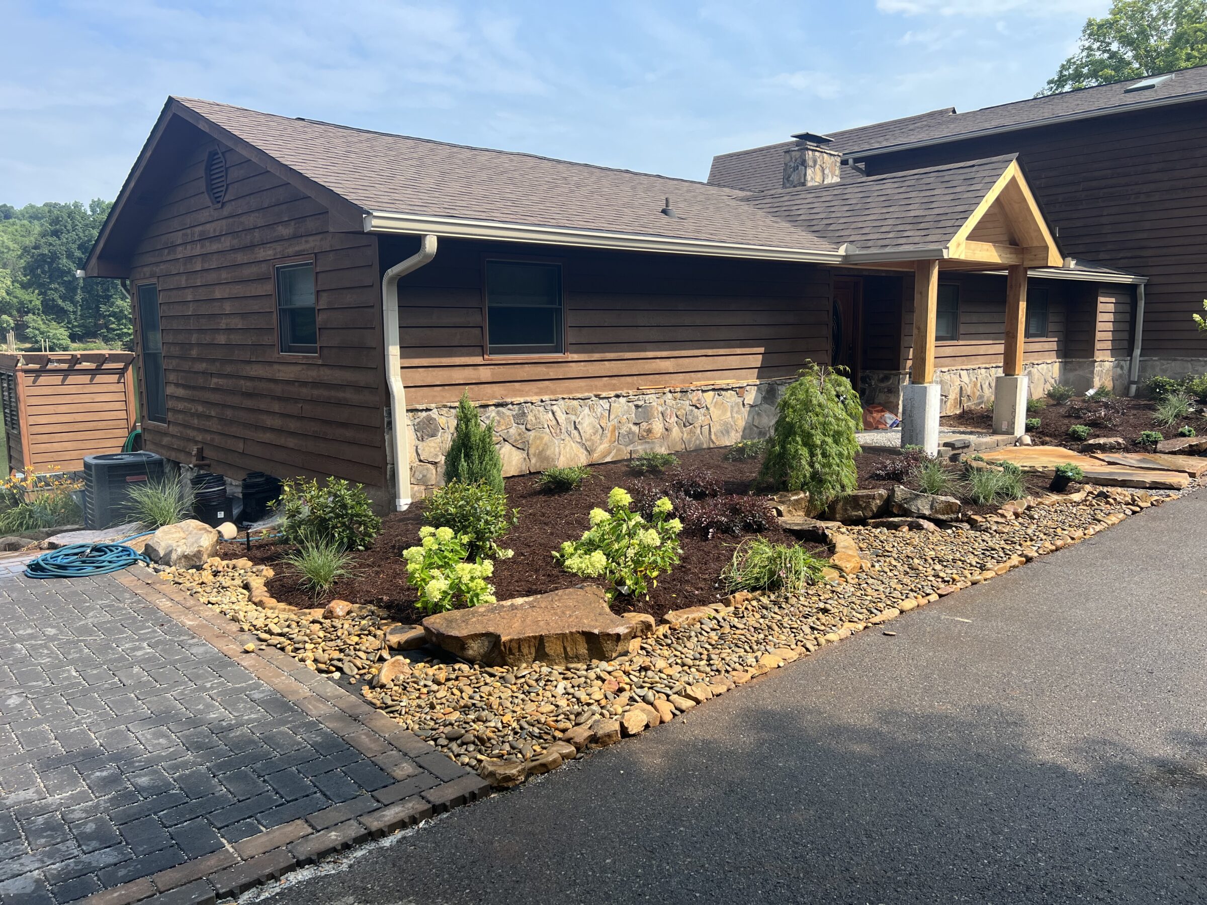 A wooden house with stone accents features a landscaped garden and paved driveway, surrounded by greenery under a clear blue sky.