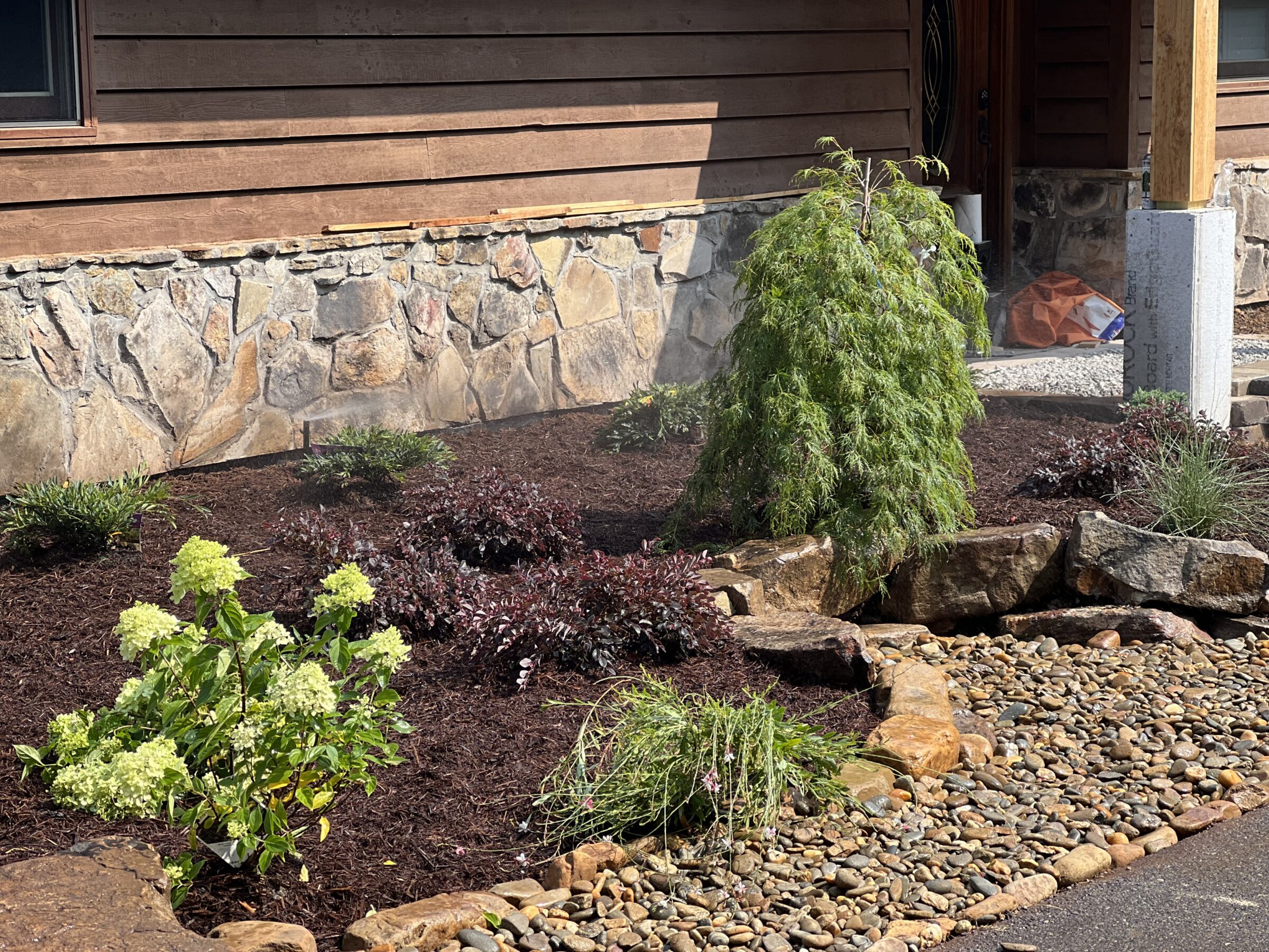 A rustic wooden house features stone landscaping with shrubs and plants. Pebble path and small tree enhance the natural aesthetic.
