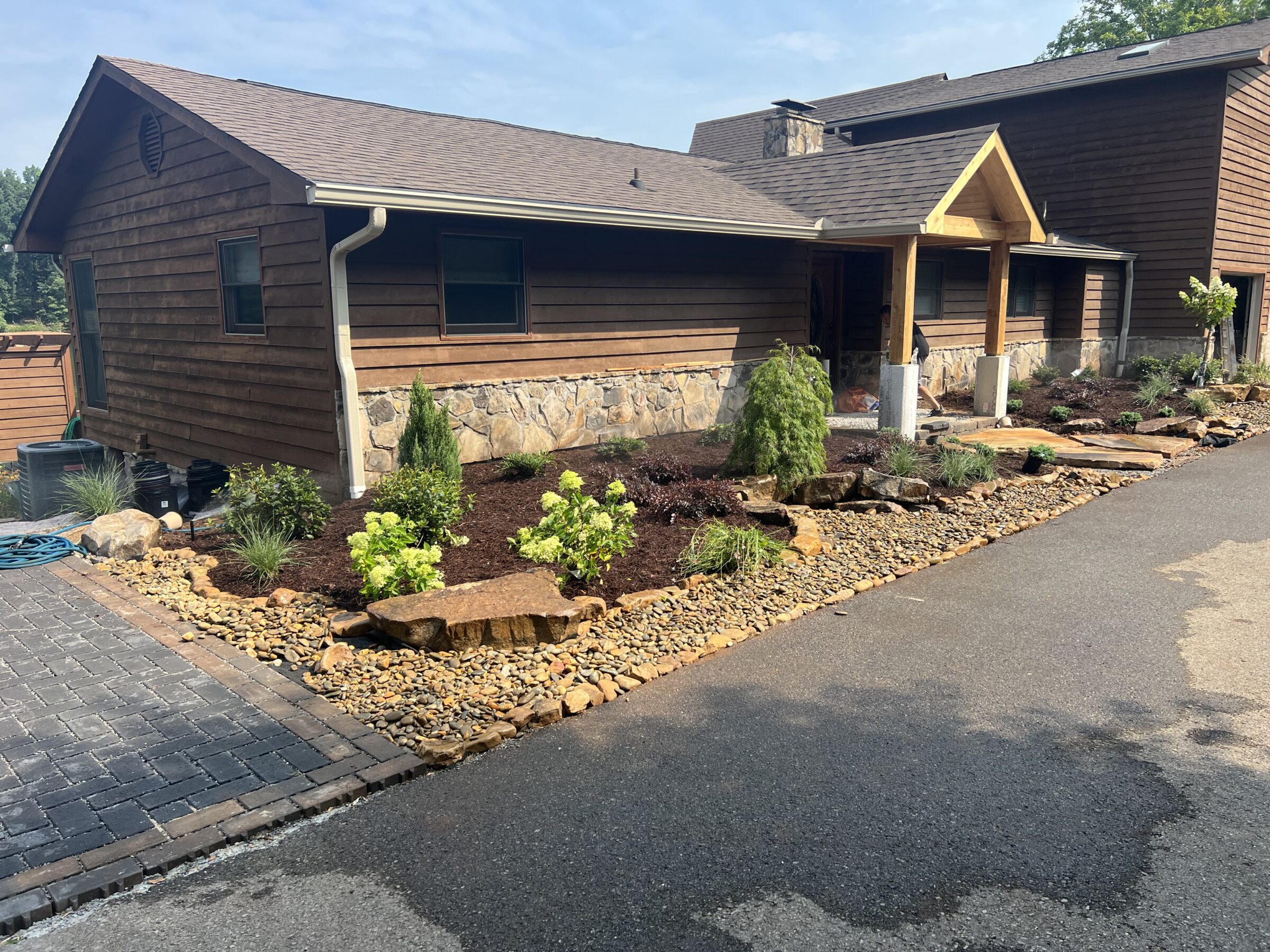 A rustic house with wooden siding and stone accents, surrounded by landscaped gardens with rocks and plants, under a clear sky.
