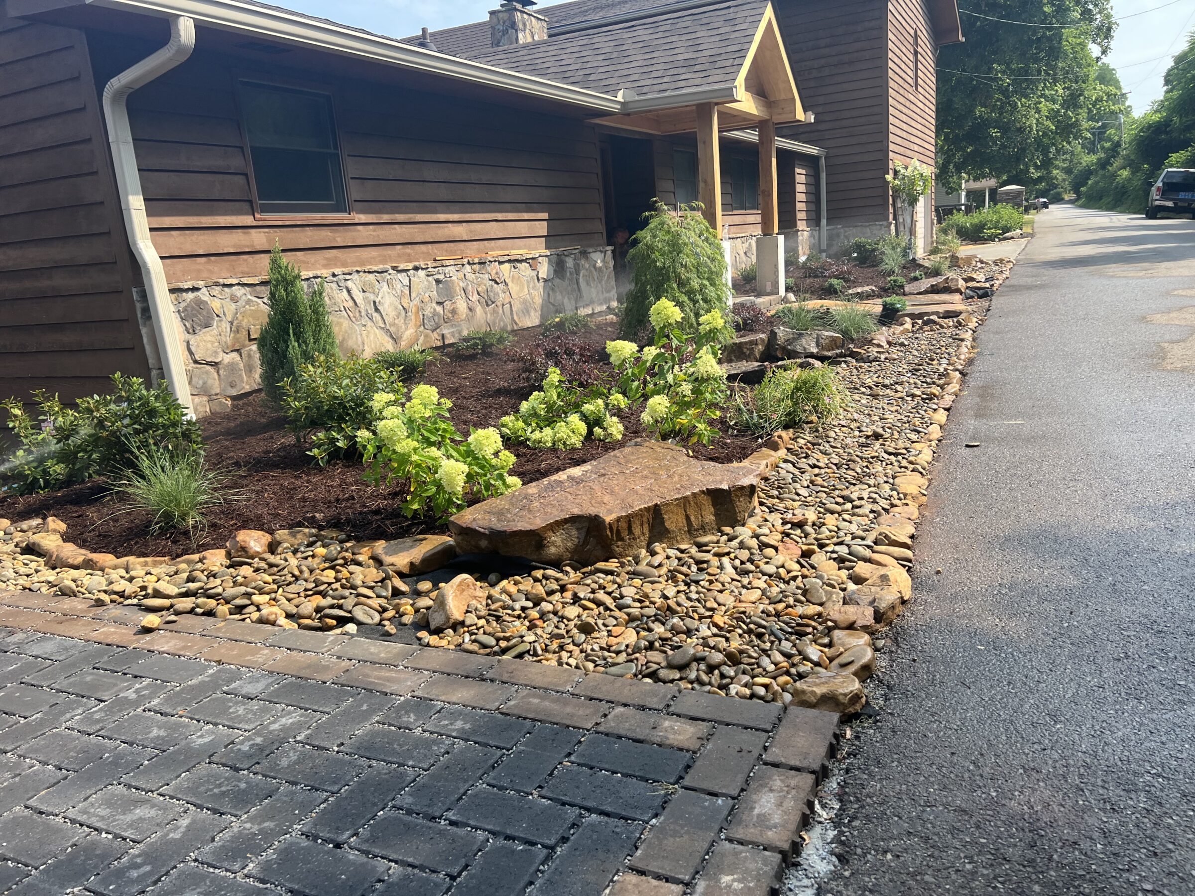 A stone and wood house with lush landscaped garden, featuring a rock border and small plants, lines a quiet residential street.