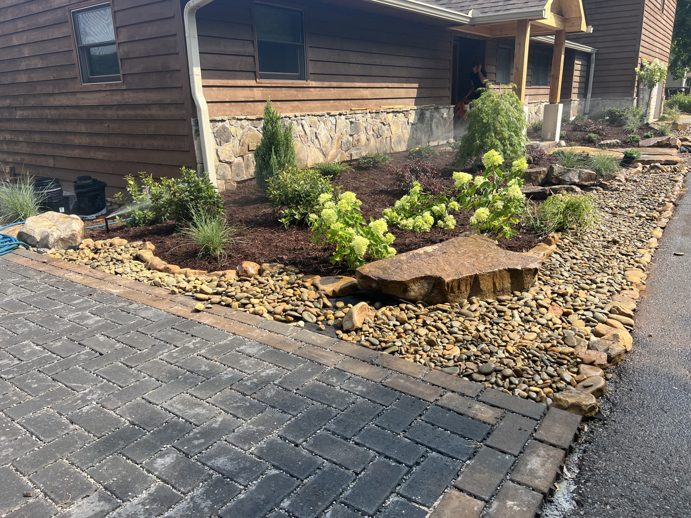 Wooden house with stone landscaping, featuring green shrubs and rocks. Brick pathway borders driveway. Person partially visible by the open door.