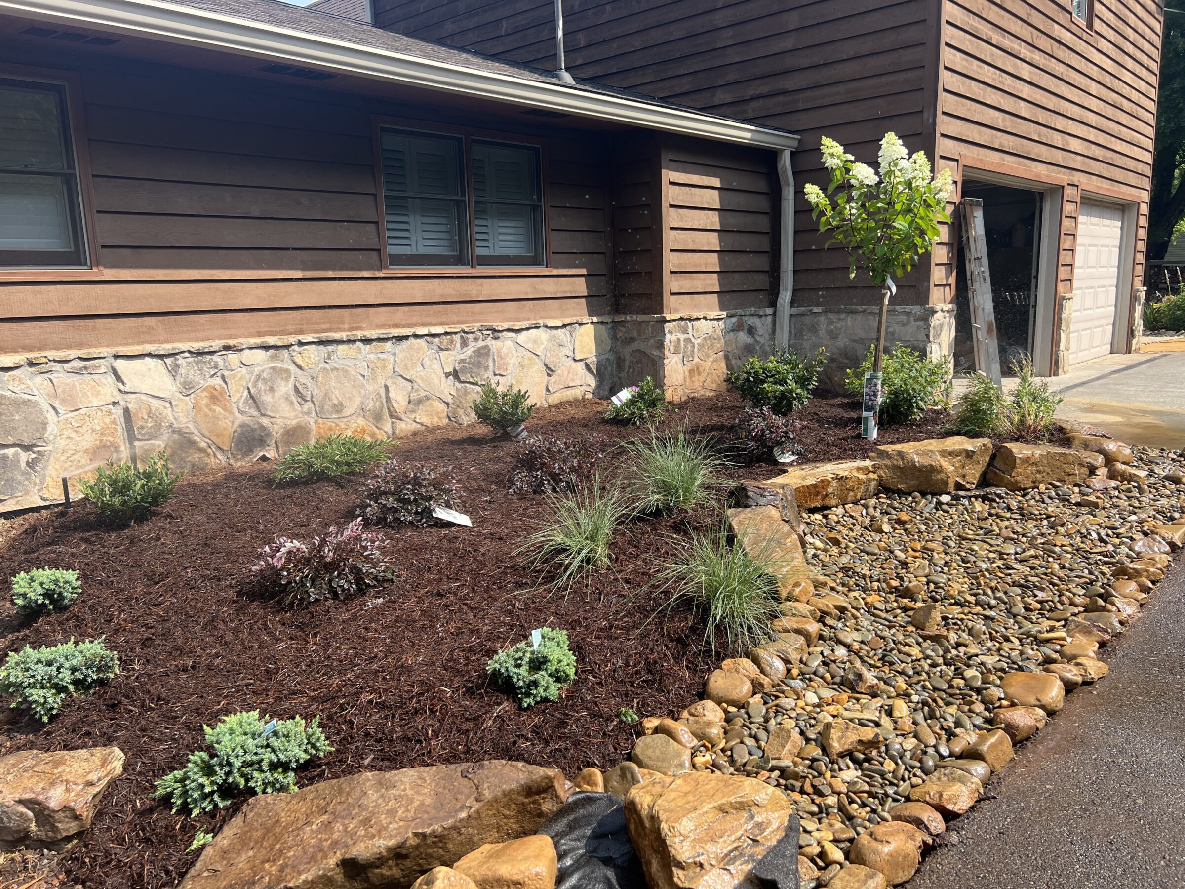 A house with stone facade features a newly landscaped yard, including mulch, small shrubs, and a pebble-lined area beside the driveway.