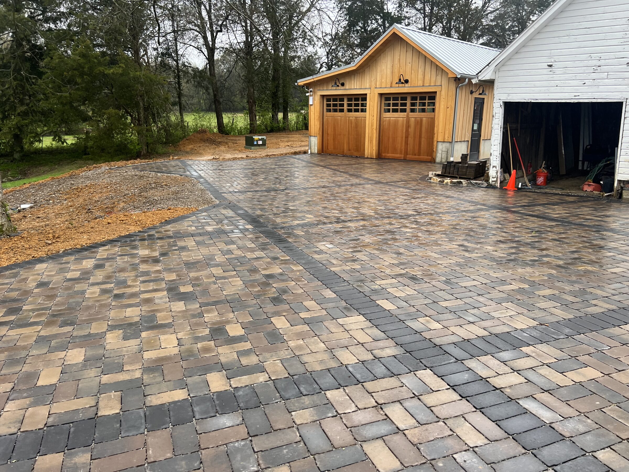 A spacious driveway with interlocking pavers leads to a wooden double garage. Surrounded by trees, it has an orderly, rustic appearance.