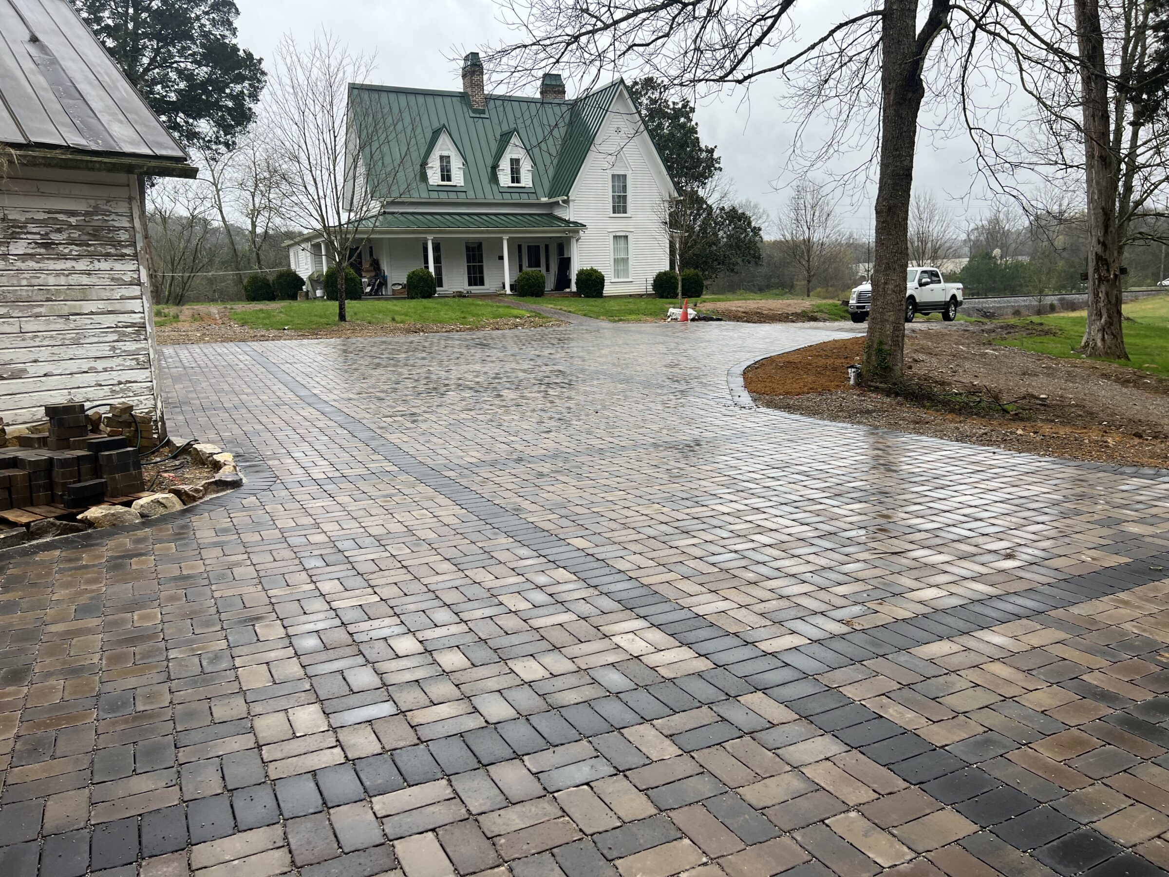 A wet brick driveway leads to a white house with a green roof and gables, surrounded by trees and a parked vehicle.