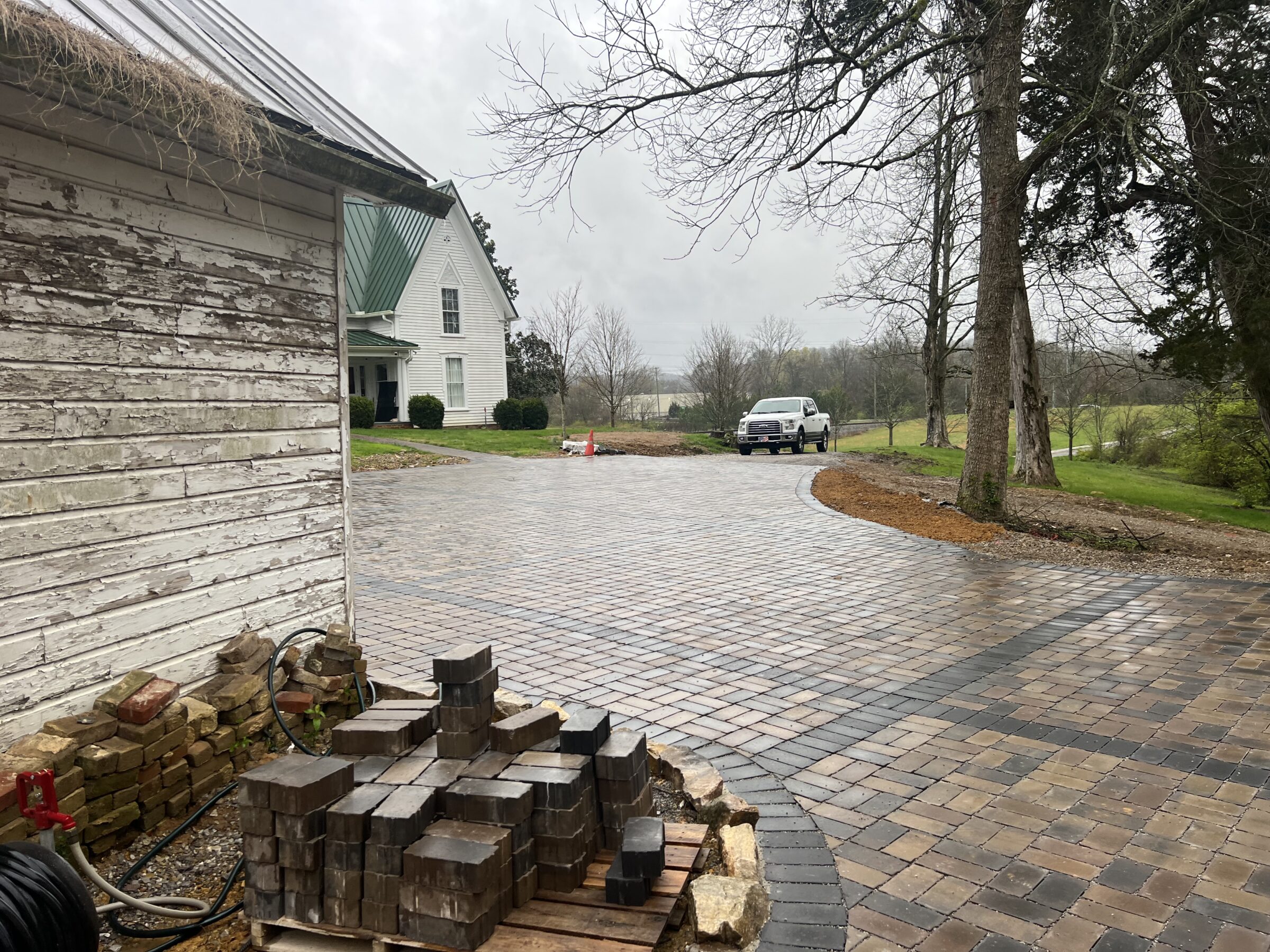 A rainy scene shows a white house with green roof, cobblestone driveway, stacked bricks, and a parked truck, surrounded by trees and grass.