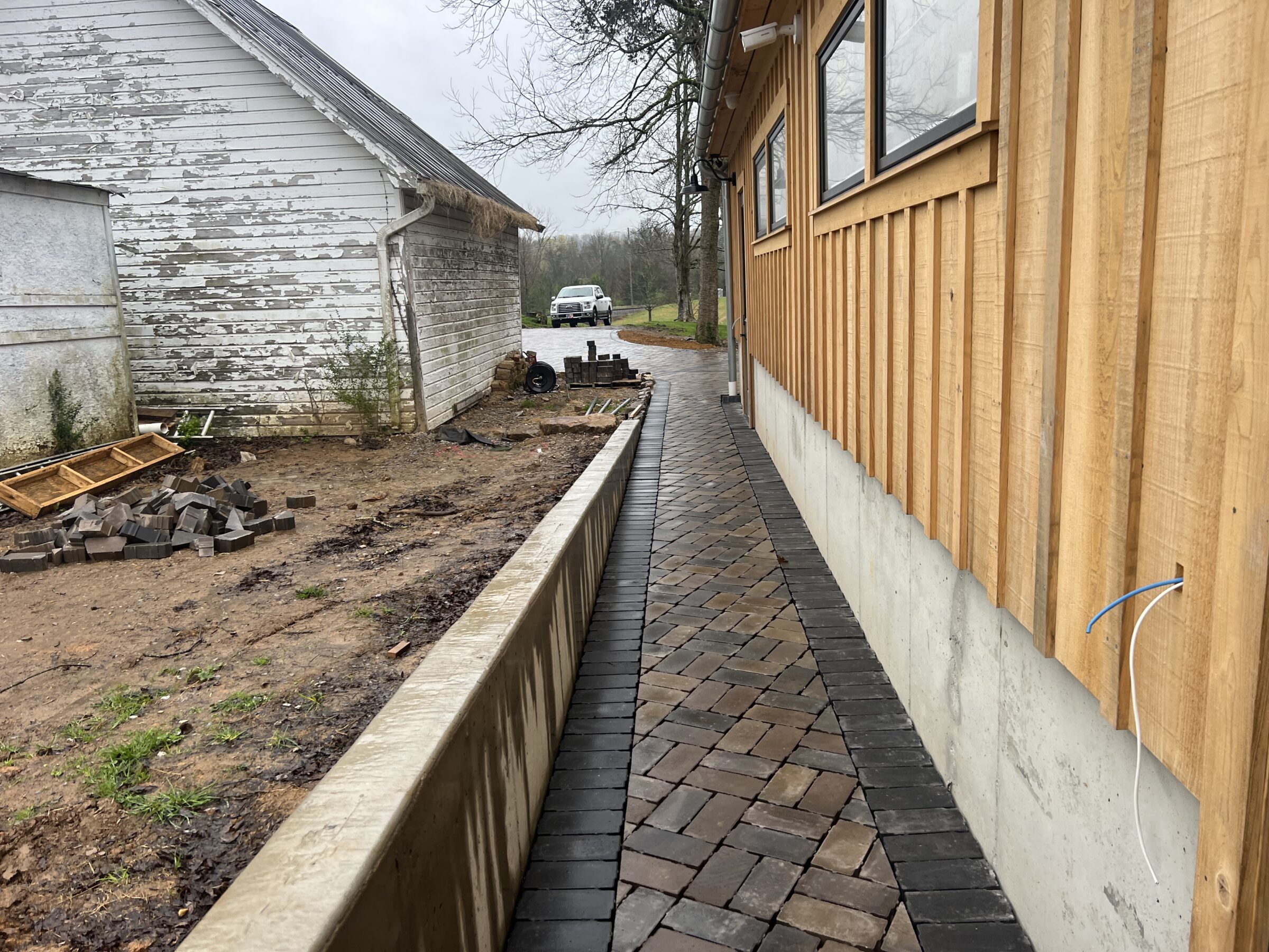 Narrow paved path between weathered white shed and wooden building, with scattered bricks and distant parked car on a cloudy day.