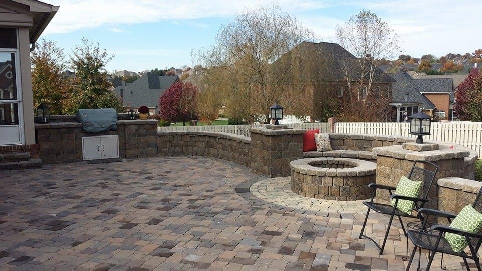 A spacious backyard patio with a fire pit, chairs, and paver flooring, surrounded by trees and residential houses in the background.