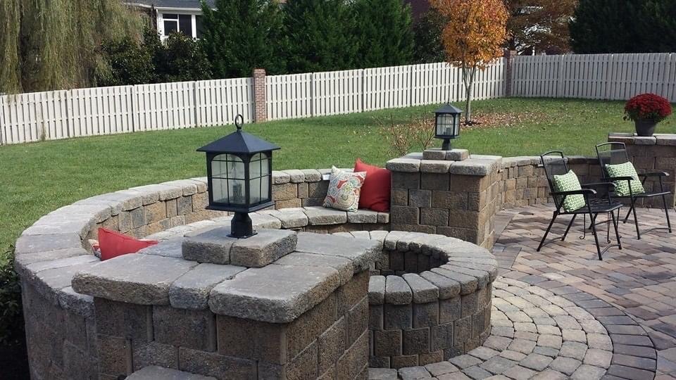 A cozy backyard patio features stone seating, colorful cushions, and lanterns, surrounded by a neatly fenced lawn and trees, creating a tranquil ambiance.