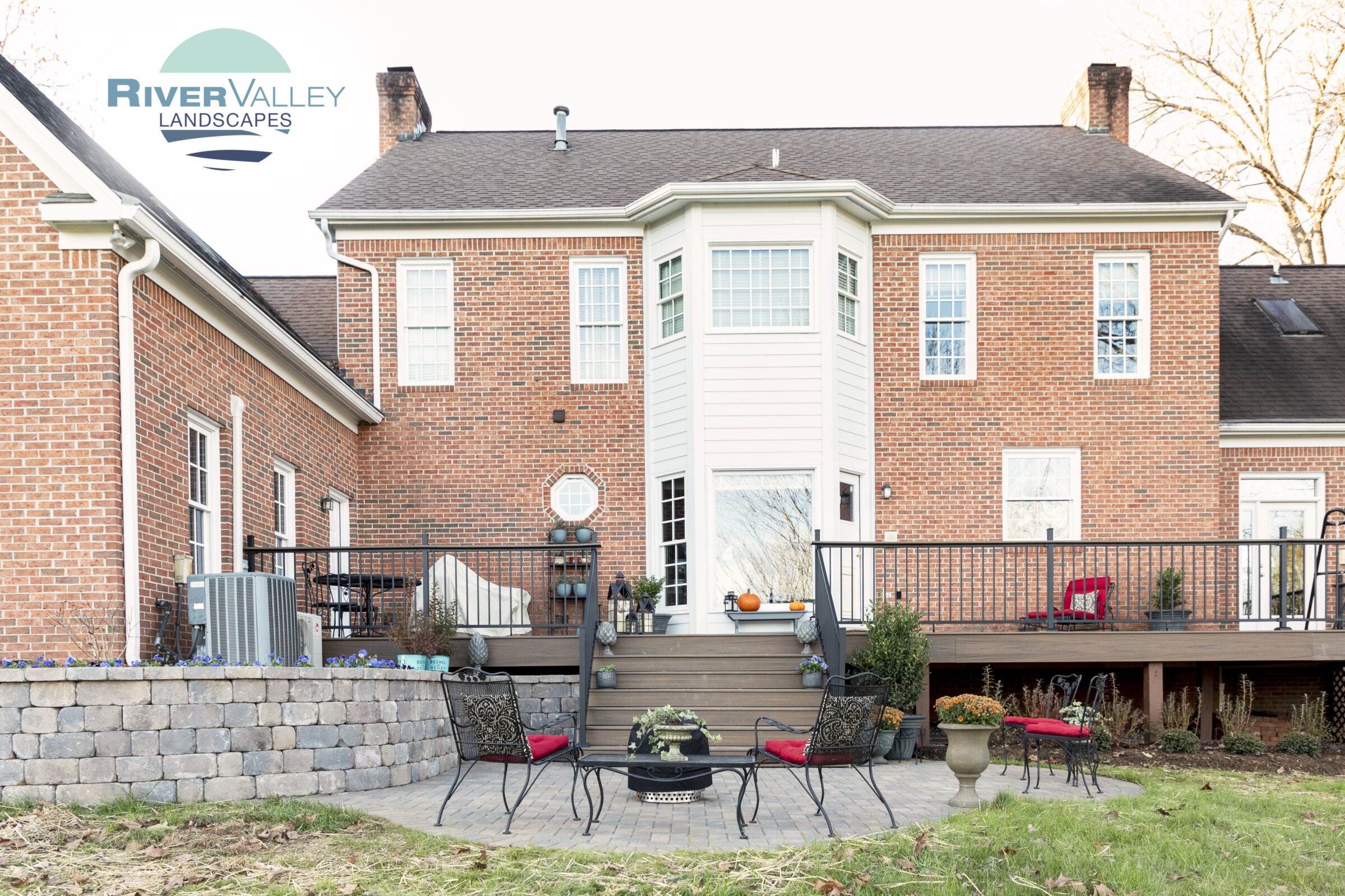 A brick house features a landscaped patio with wrought iron furniture and plants. A sign reads "River Valley Landscapes" in the top left.
