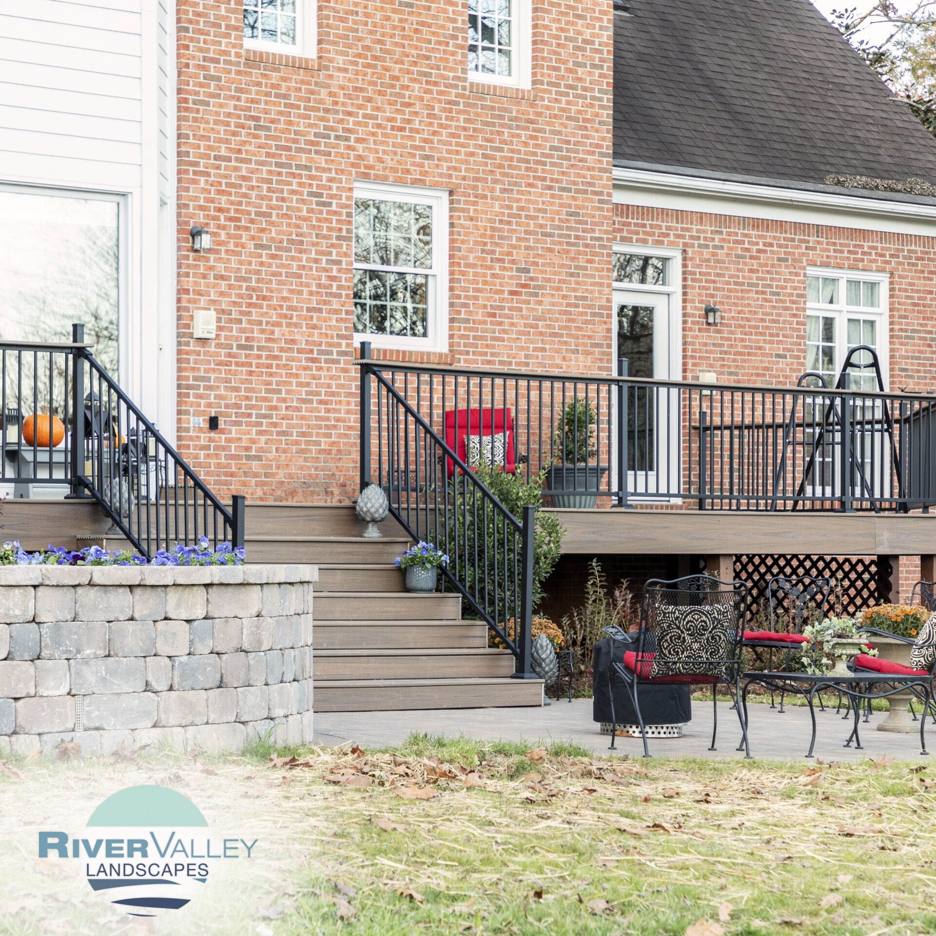 A brick house features a patio with black railings, outdoor furniture, potted plants, and a pumpkin decoration, creating a cozy, landscaped setting.